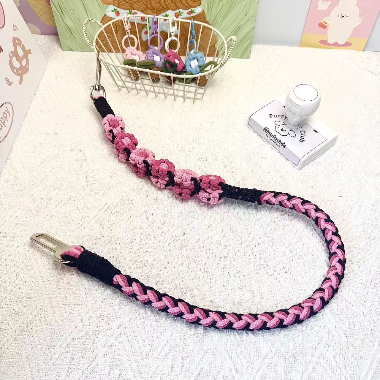 A handmade dog car safety belt in black and pink with floral accents, featuring a metal clasp and a buckle, placed on a textured white fabric background.