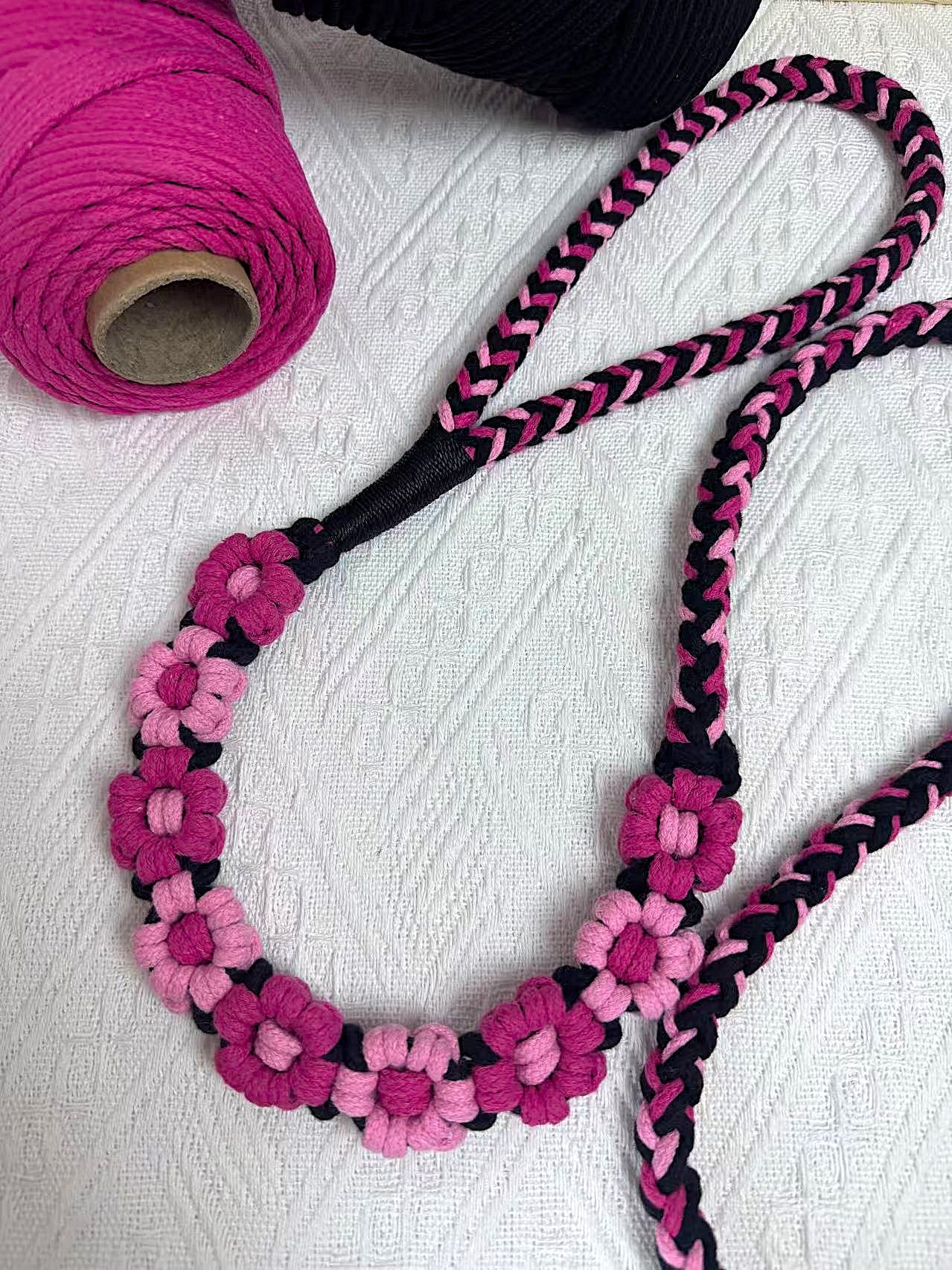 A detailed view of a black and pink handwoven dog leash with a charming floral design. The leash is elegantly placed on a textured white surface, highlighting its intricate craftsmanship and vibrant colors.