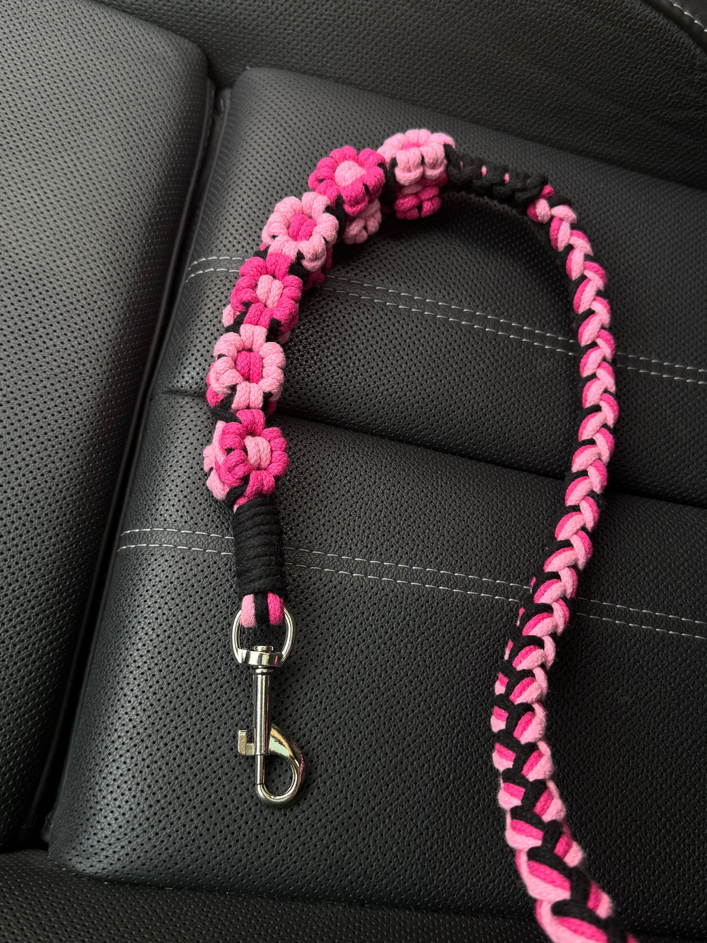 A black and pink handwoven dog safety seatbelt placed on a car seat. The design showcases intricate floral patterns and a secure metal clip, blending functionality with elegance for your pet's travels.