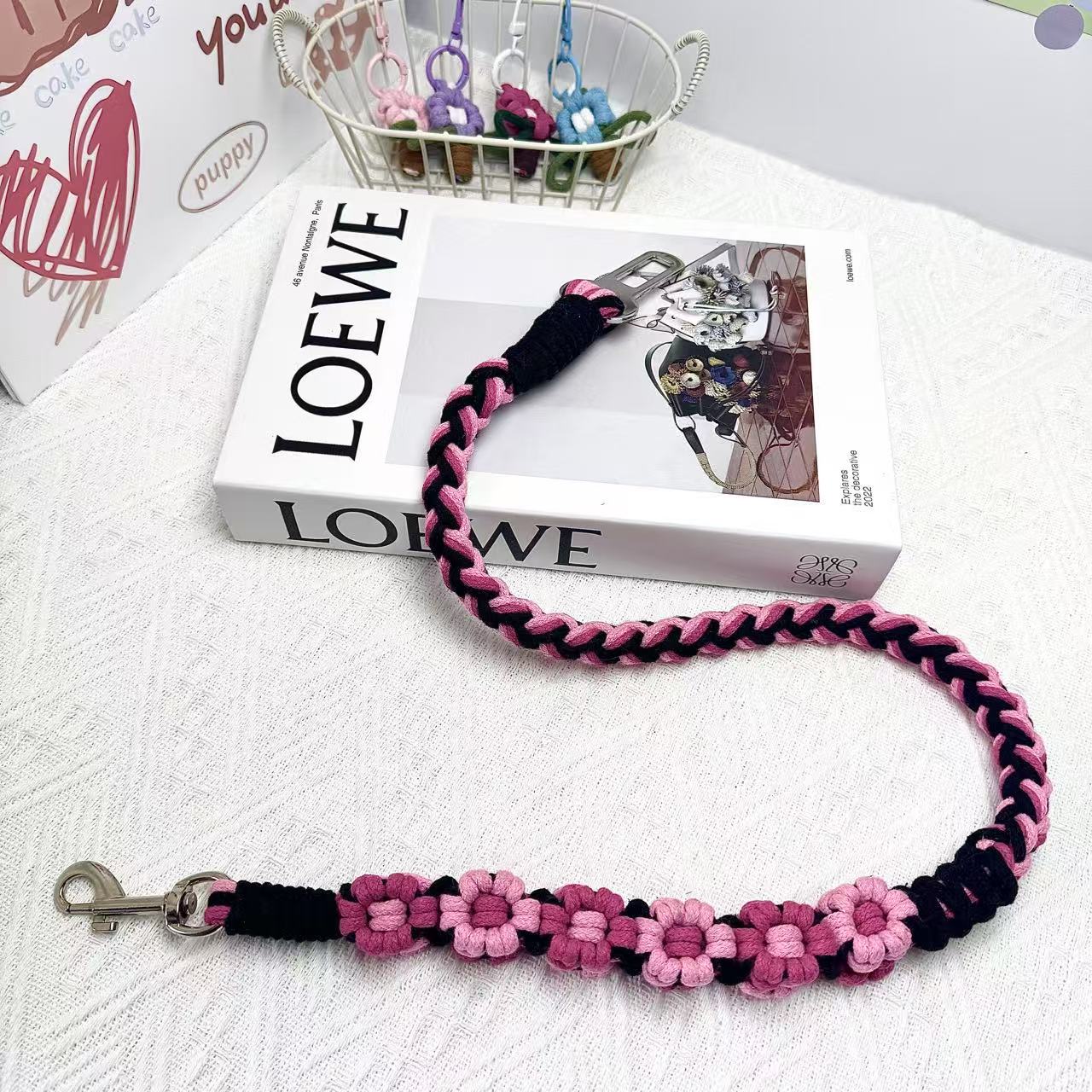 A handwoven black and pink dog safety seatbelt displayed stylishly on a textured surface with floral details and a secure clasp, adding charm to pet accessories.