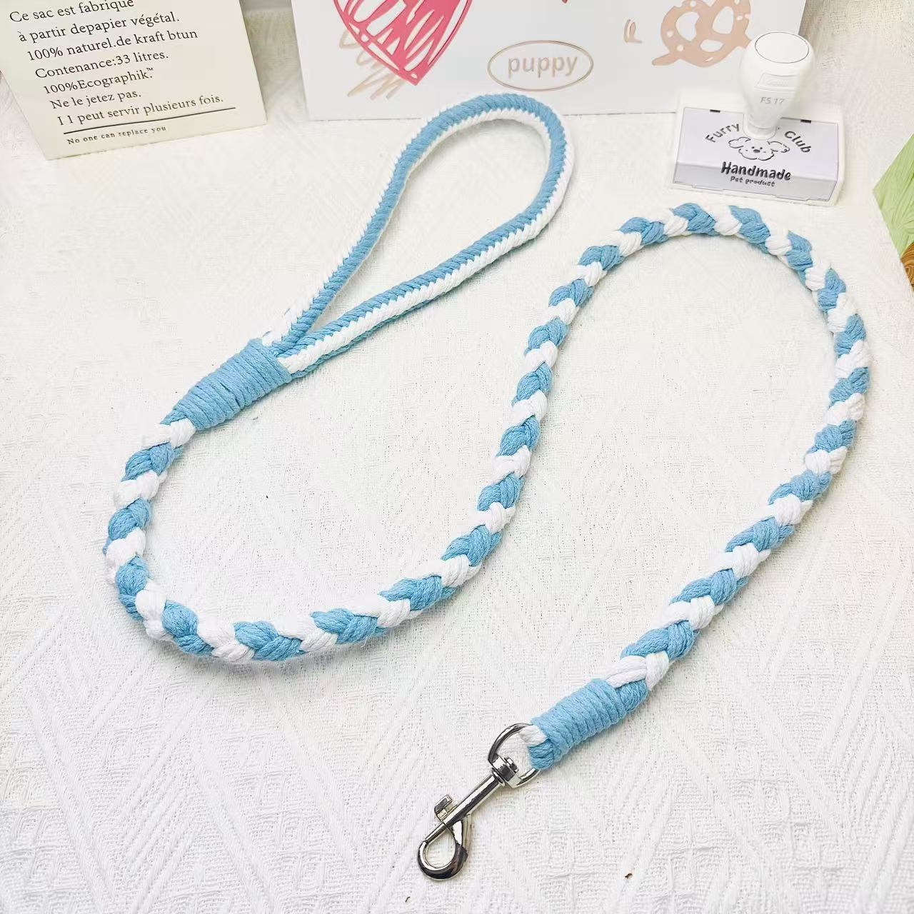 A handmade dog leash in blue and white, featuring a metal clasp, placed on a textured white fabric background.