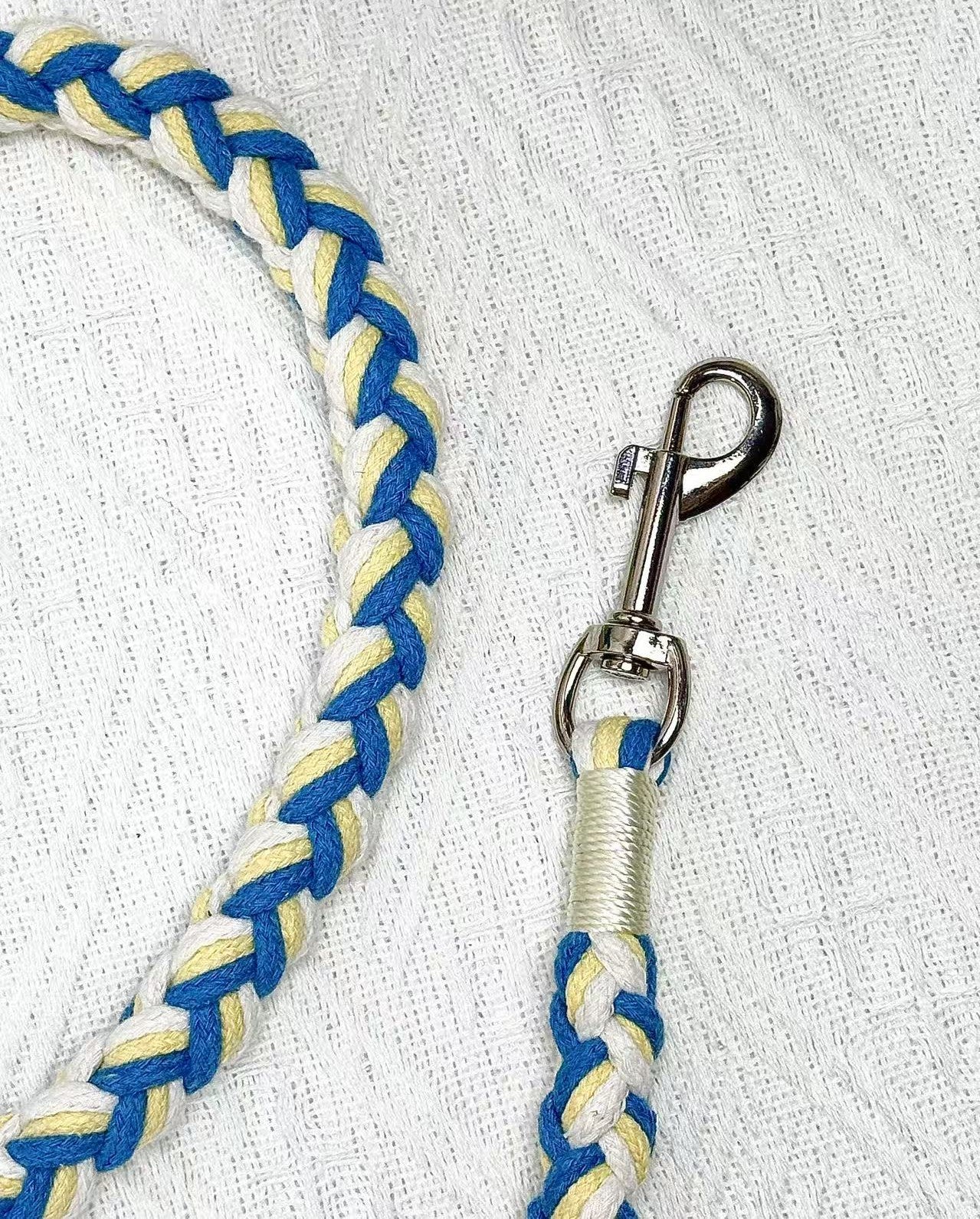 A close-up of a blue, yellow, and white handwoven dog leash, showcasing the durable braided design and a polished metal clip for secure attachment.