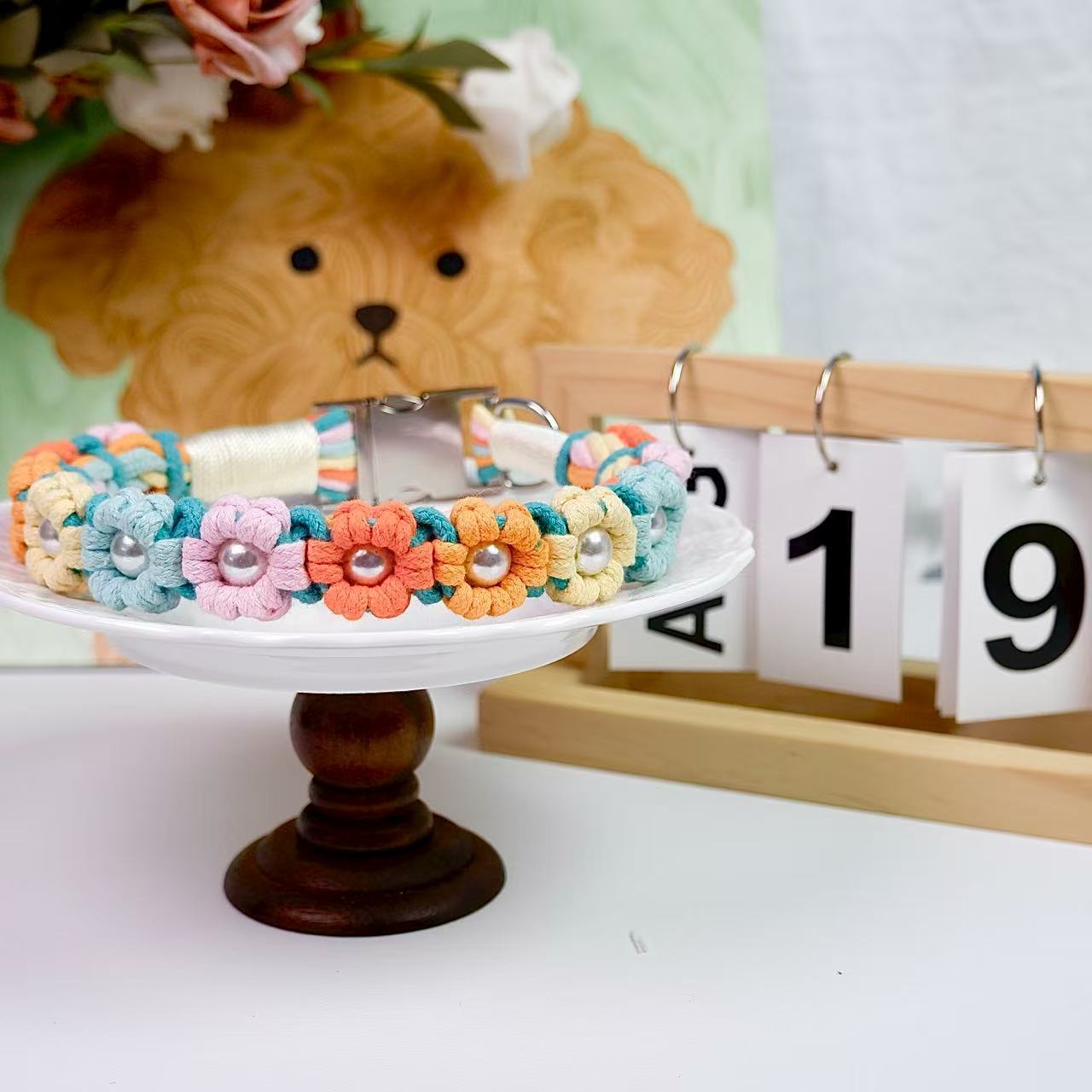 Handmade dog collar with multicolor flower knots and pearl centers, displayed on a white stand with a wooden base.