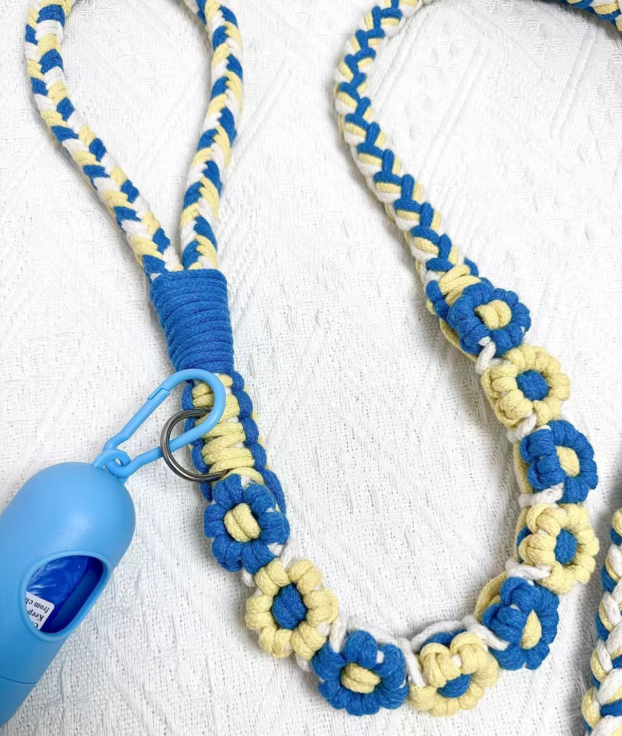 Front half of a blue and yellow handwoven dog leash with a handle, flower accents, and a blue waste bag holder, set on a white background.