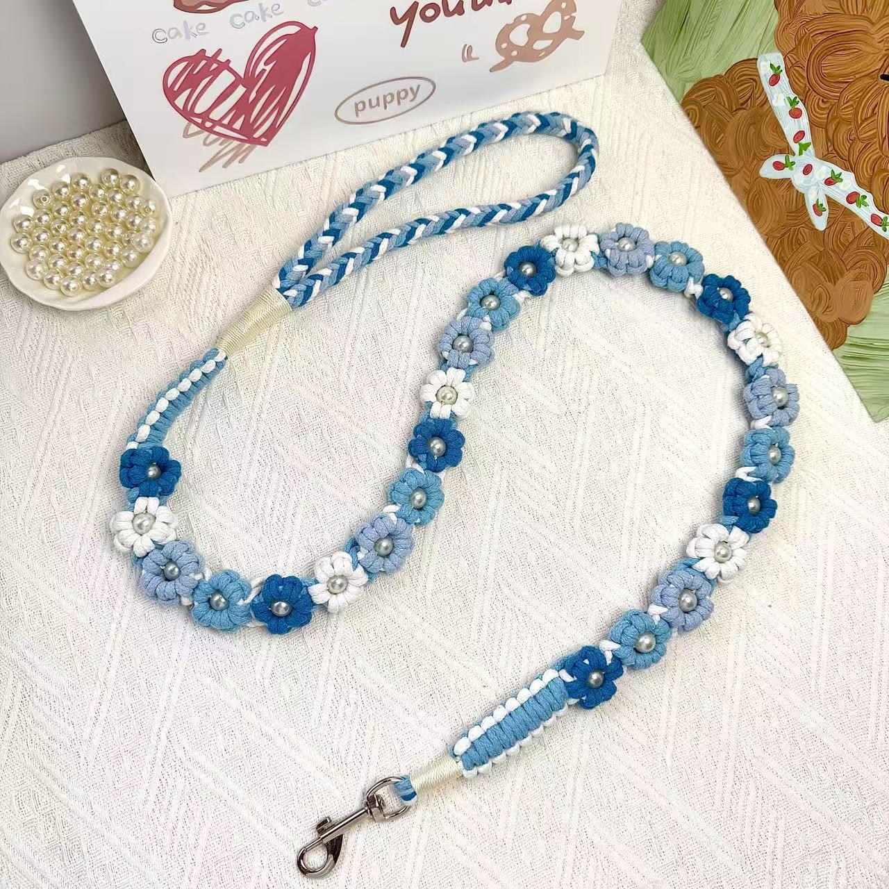A handmade dog leash in gradient blue with floral accents and pearl details, featuring a metal clasp, placed on a textured white fabric background.