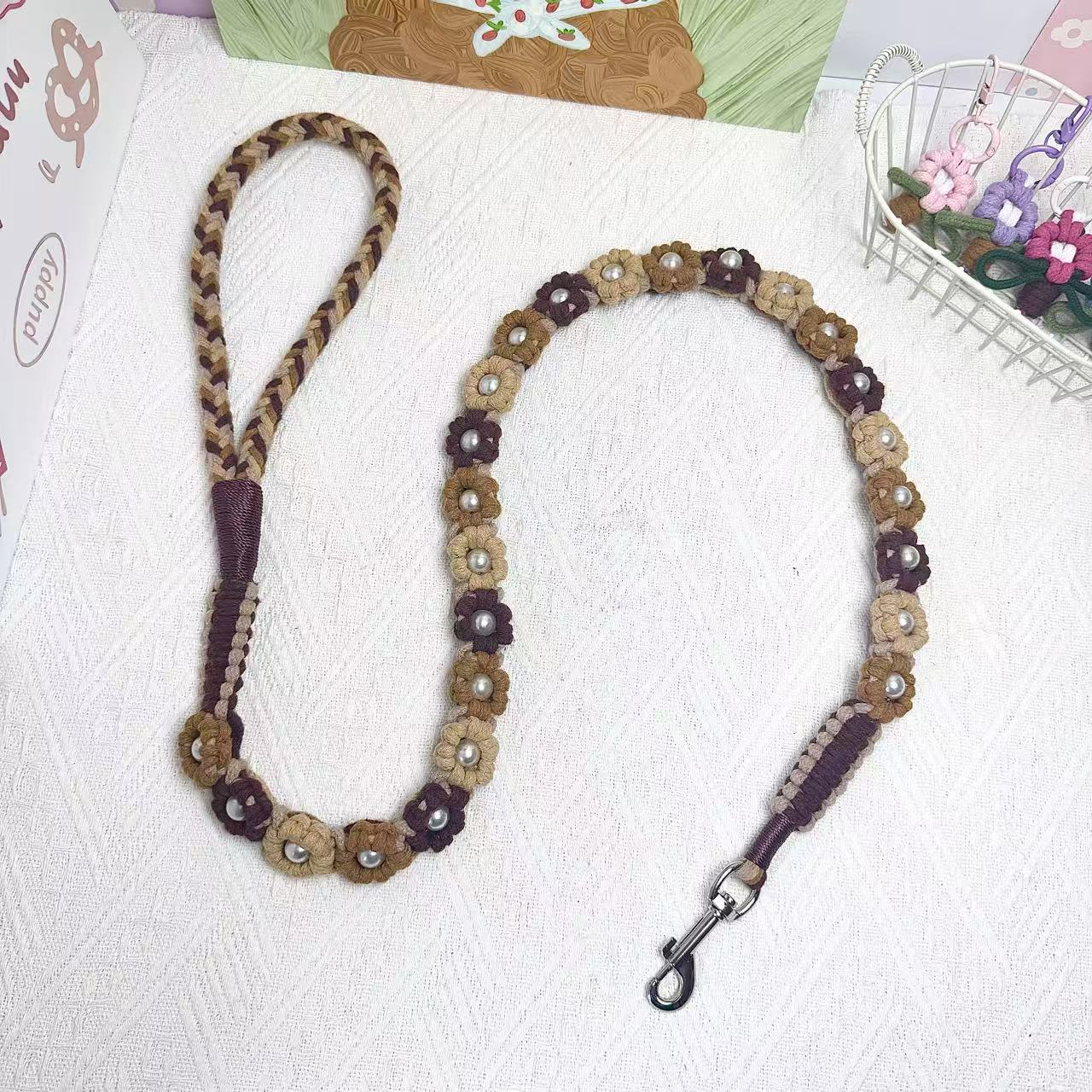 A handmade dog leash in gradient brown with floral accents and pearl details, featuring a metal clasp, placed on a textured white fabric background.