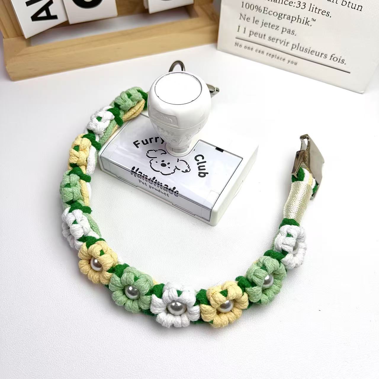 Handwoven grass green dog collar with colorful flowers and pearl accents, featuring a stainless steel clasp and D-ring, placed by a white stamp.