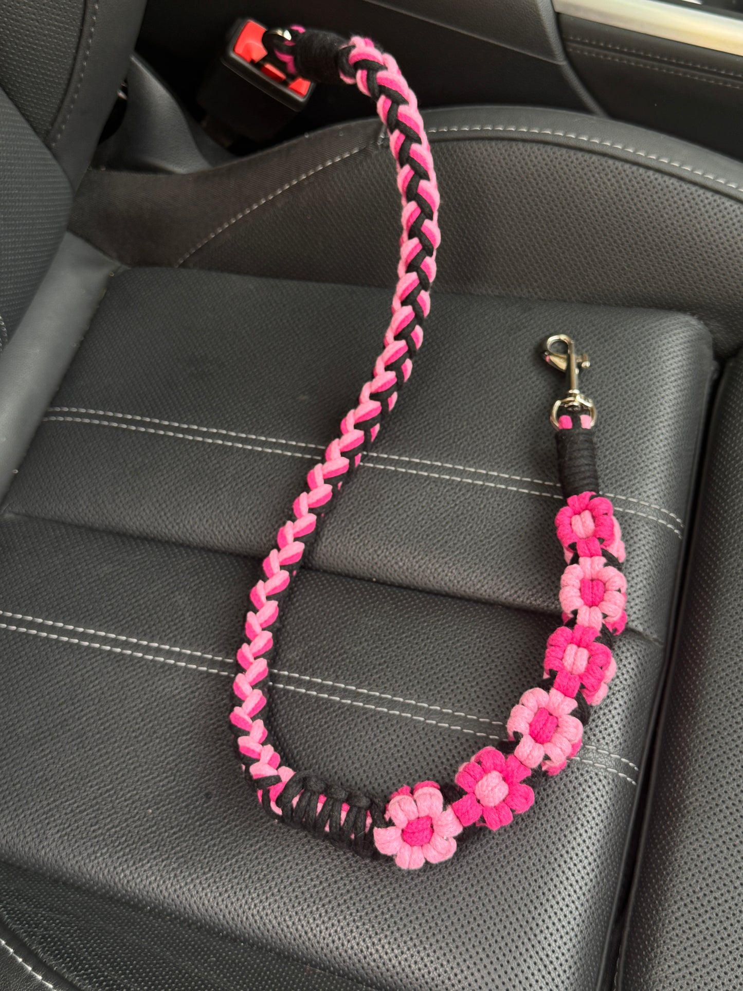 A handcrafted dog seatbelt featuring a pink floral design, intricately braided with soft pastel tone and elegant black tone. The seatbelt is placed on a black car seat, showcasing its elegant floral accents and sturdy clasp attachment.