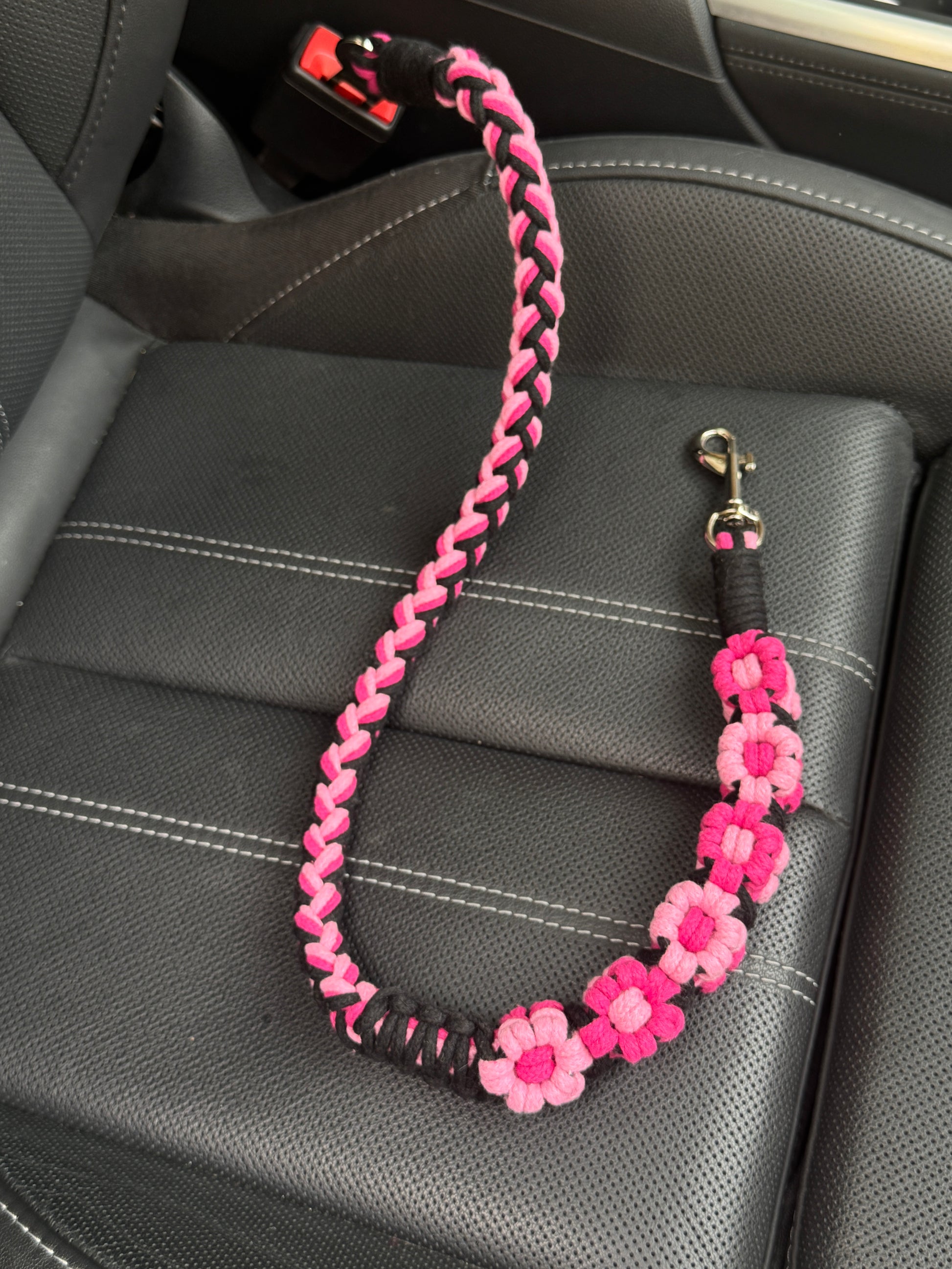A handcrafted dog seatbelt featuring a pink floral design, intricately braided with soft pastel tone and elegant black tone. The seatbelt is placed on a black car seat, showcasing its elegant floral accents and sturdy clasp attachment.