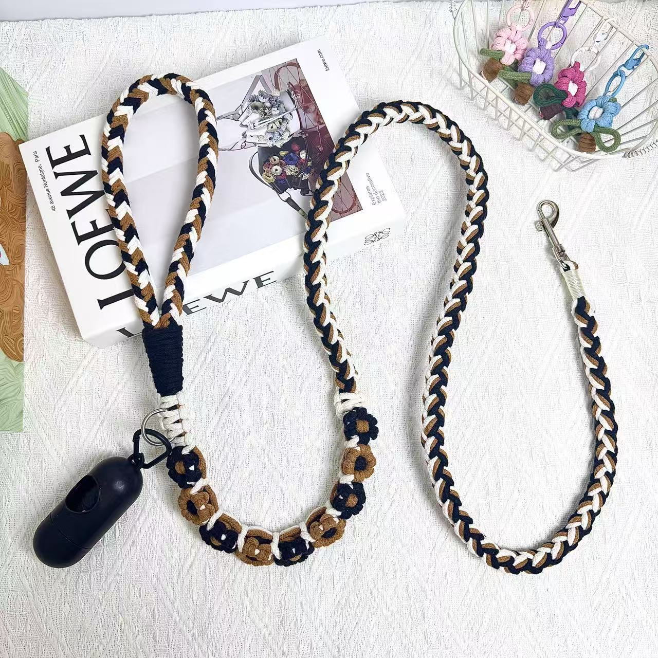 A beautifully handwoven black and brown dog leash featuring a sturdy loop handle, a metal clip for secure attachment, and an integrated waste bag holder, displayed on a white surface with decorative elements.
