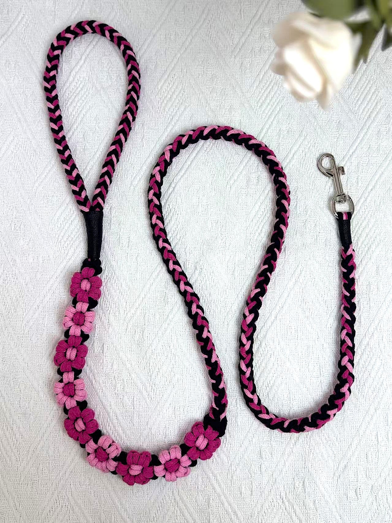 A beautifully handwoven black and pink dog leash featuring a sturdy loop handle and a metal clip for secure attachment, displayed on a white surface with decorative elements.