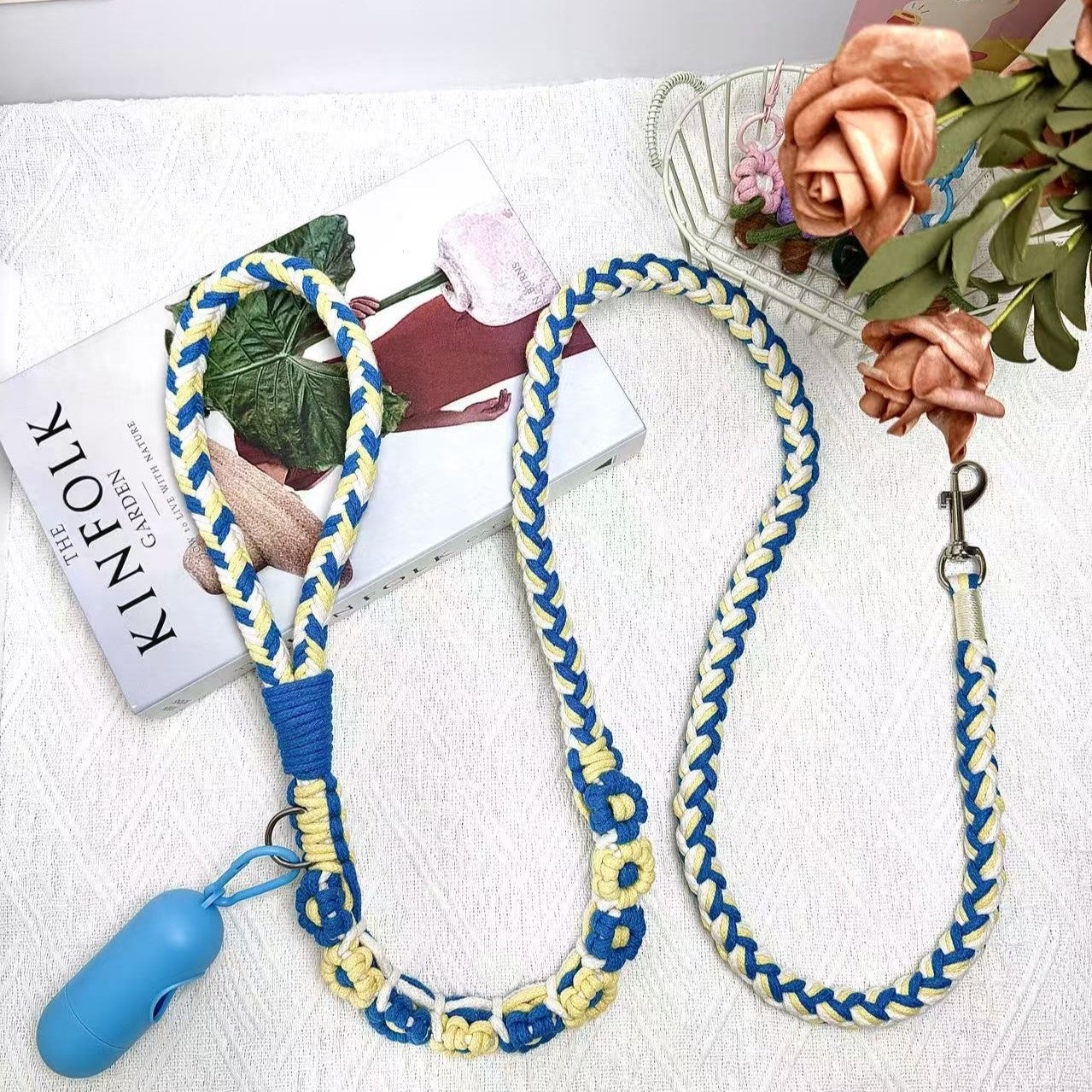 A beautifully handwoven blue and yellow dog leash featuring a sturdy loop handle, a metal clip for secure attachment, and an integrated waste bag holder, displayed on a white surface with decorative elements.