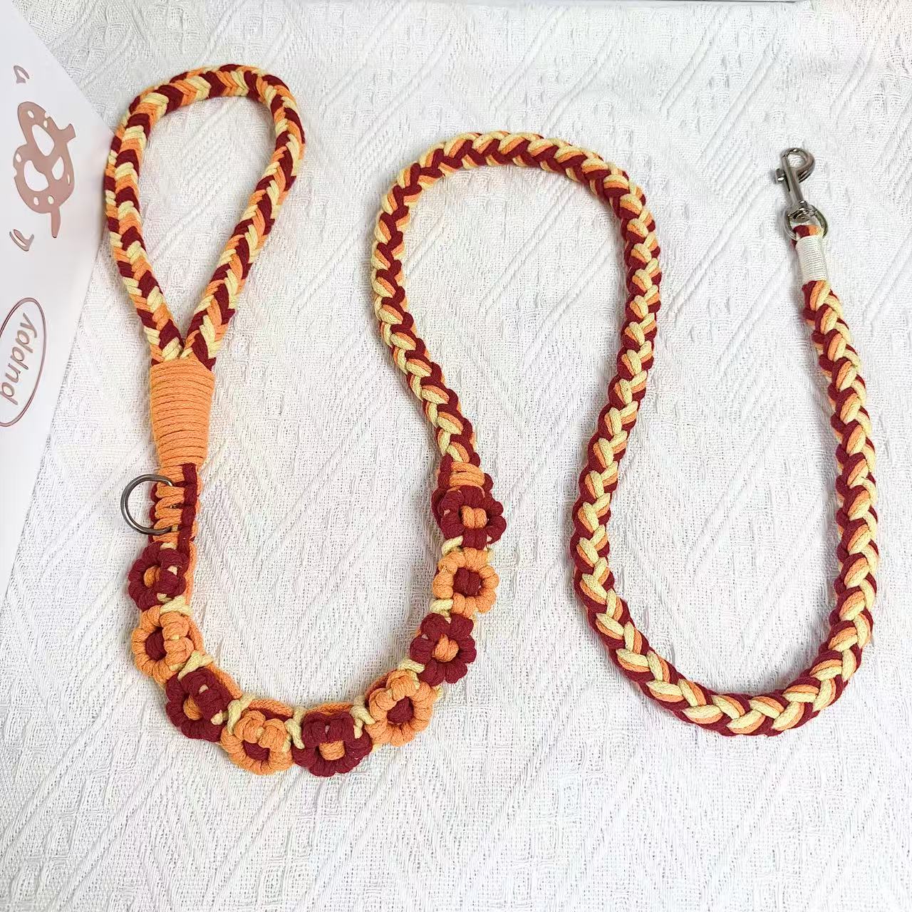 A beautifully handwoven orange and red dog leash featuring a sturdy loop handle and a metal clip for secure attachment, displayed on a white surface with decorative elements.