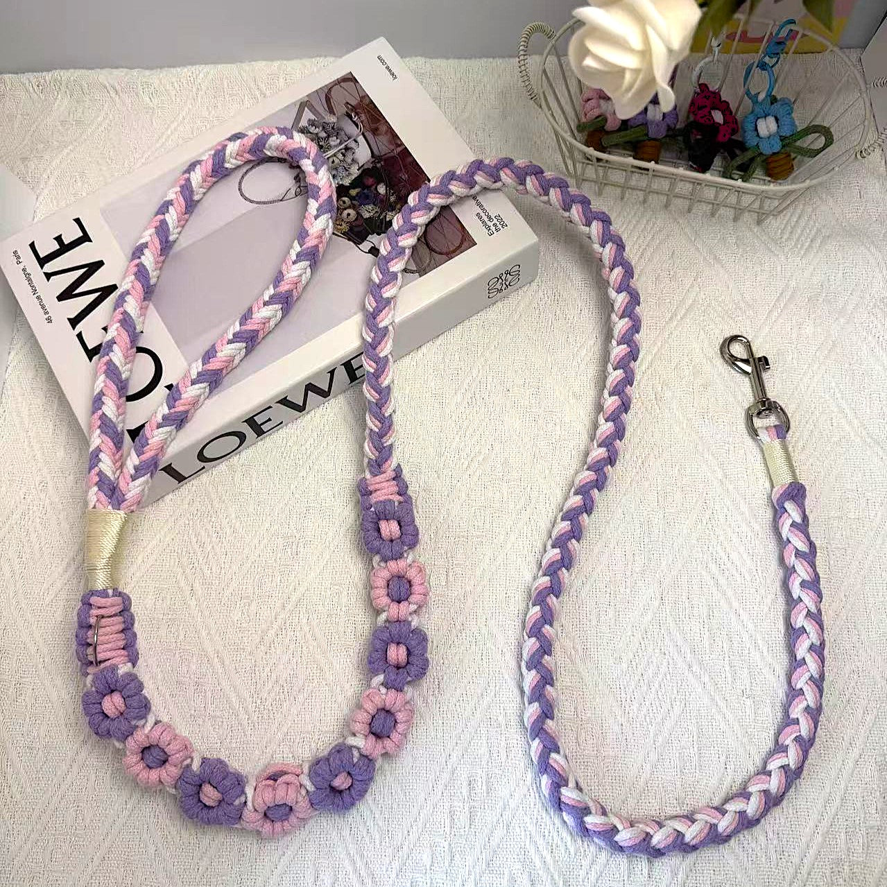 A beautifully handwoven purple and pink dog leash featuring a sturdy loop handle and a metal clip for secure attachment, displayed on a white surface with decorative elements.