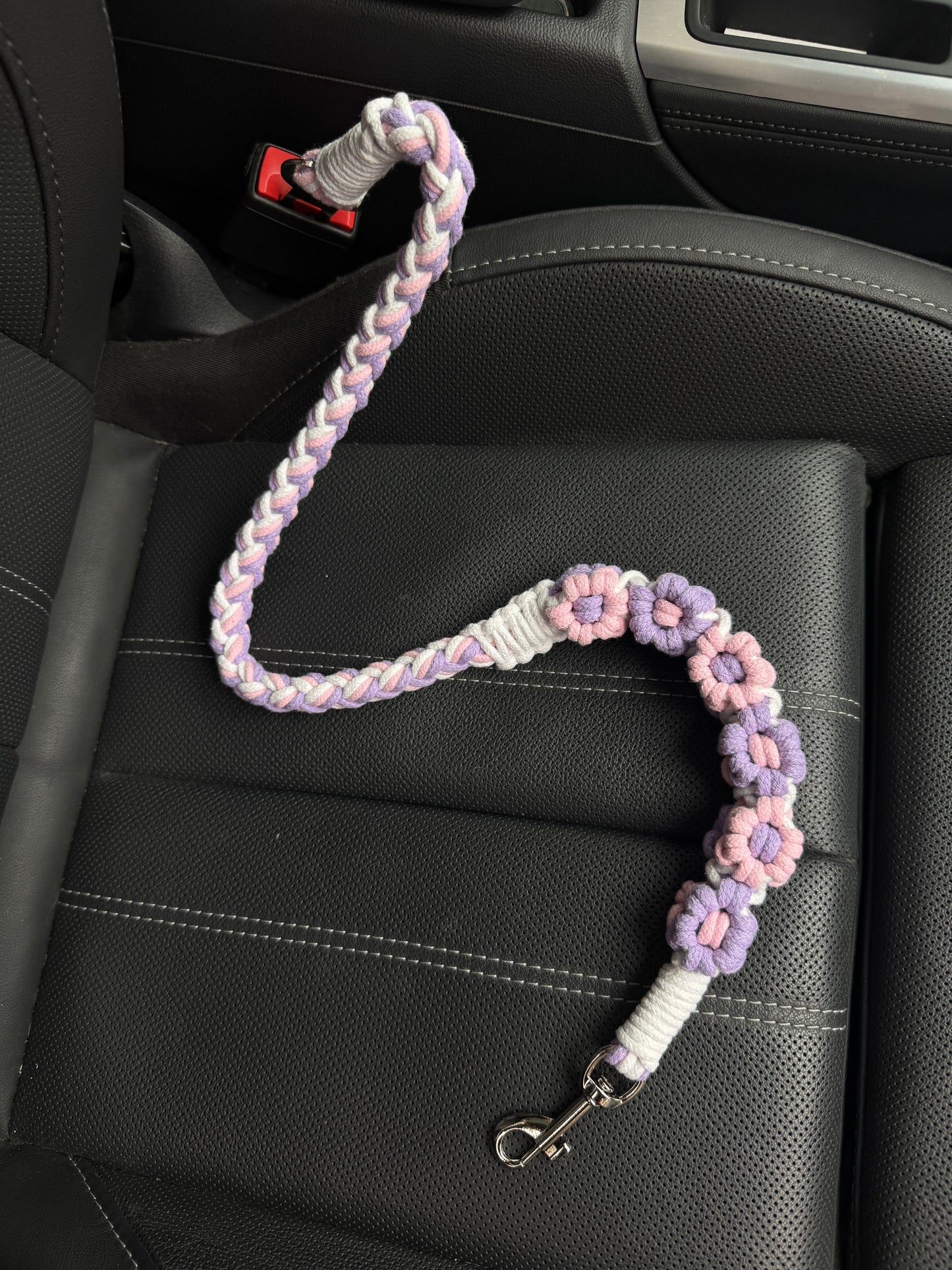 A handcrafted dog seatbelt featuring a pink and purple floral design, intricately braided with soft pastel tones. The seatbelt is placed on a black car seat, showcasing its elegant floral accents and sturdy clasp attachment.