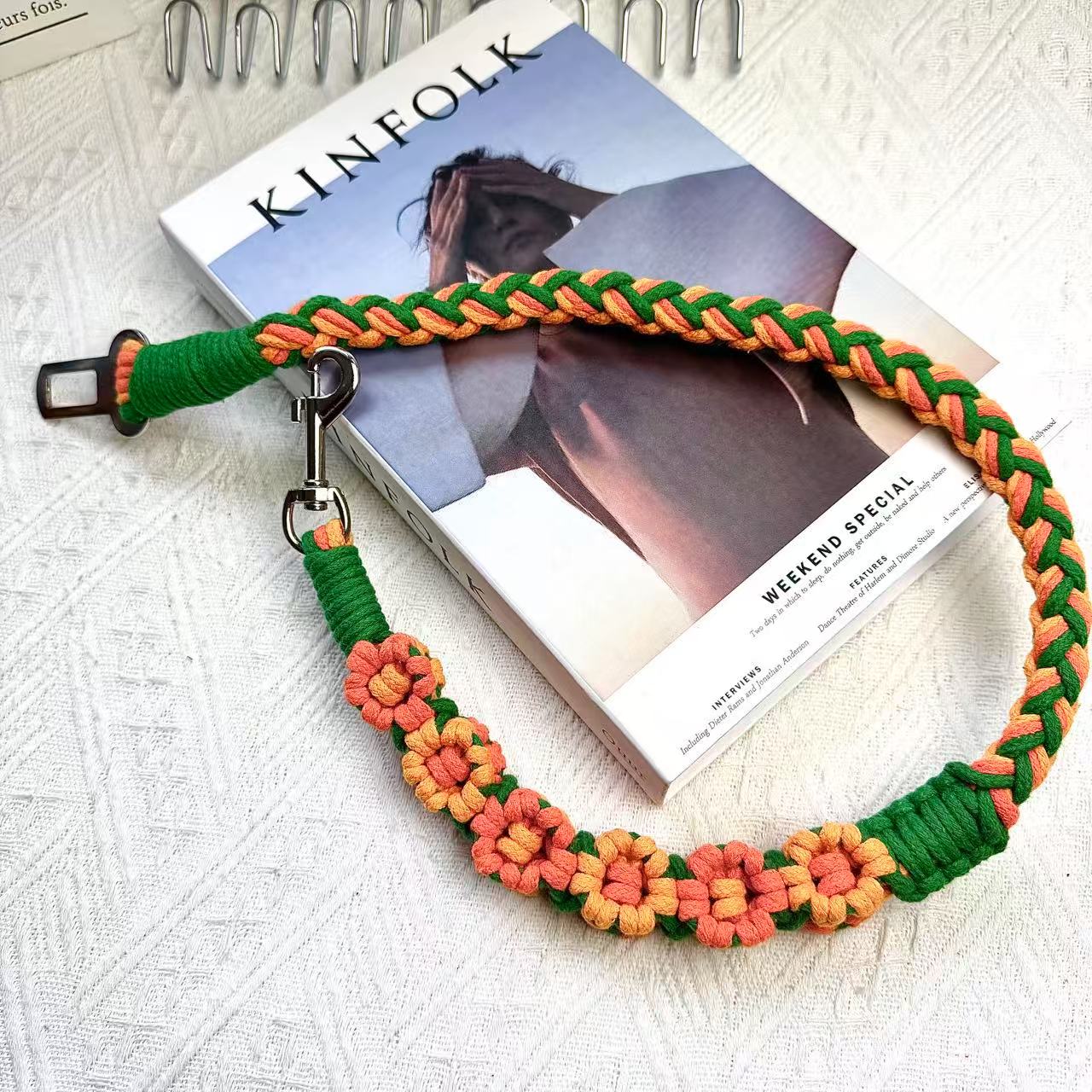 A handmade dog car safety belt in orange and green with floral accents, featuring a metal clasp and a buckle, placed on a textured white fabric background.