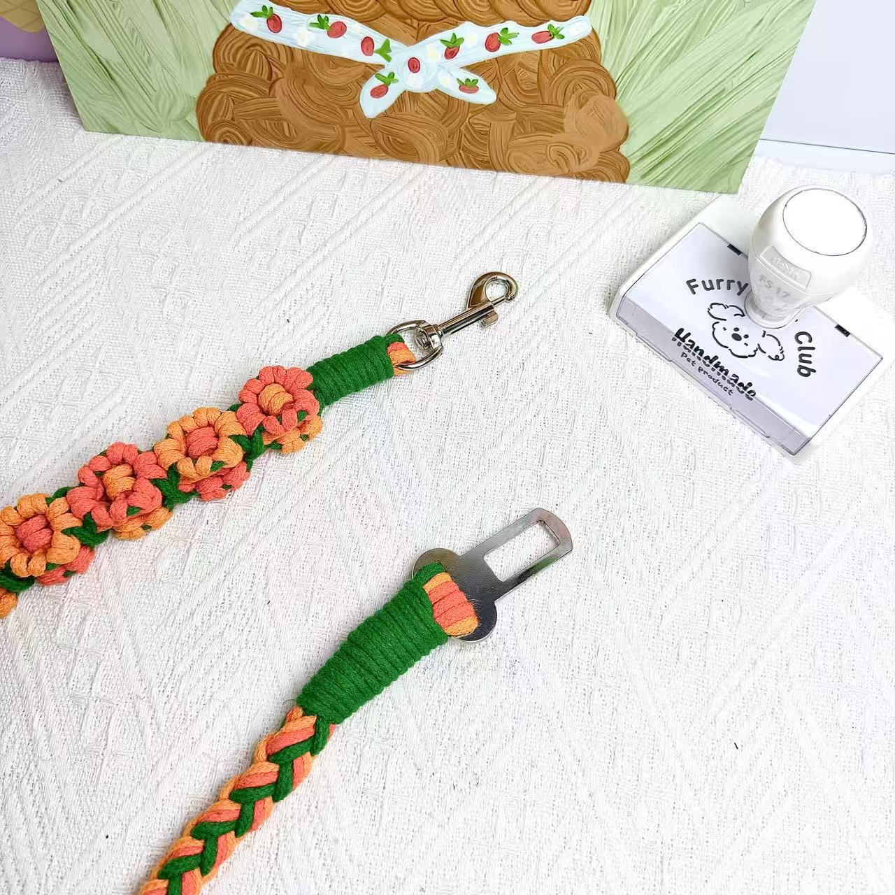 A handcrafted dog seatbelt attachment featuring a braided rope design in pastel orange and green. The sturdy metal clip is securely wrapped with green cord, ensuring durability.