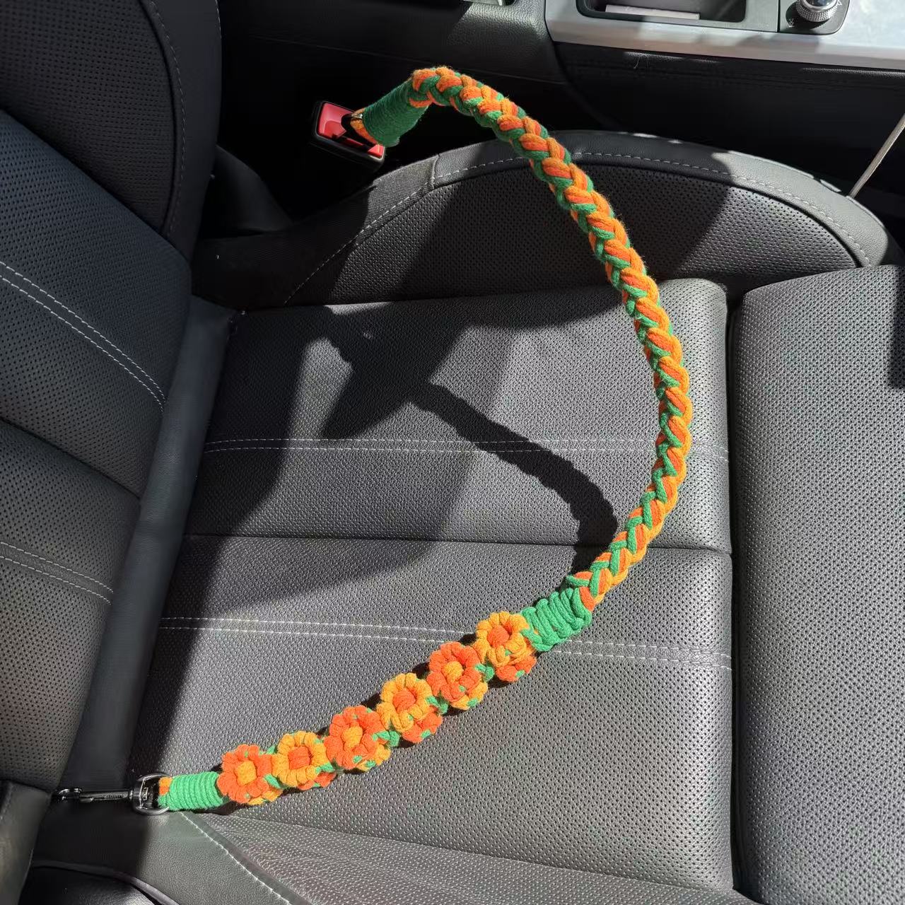 An orange and green handwoven dog safety seatbelt placed on a car seat. The design showcases intricate floral patterns and a secure metal clip, blending functionality with elegance for your pet's travels.