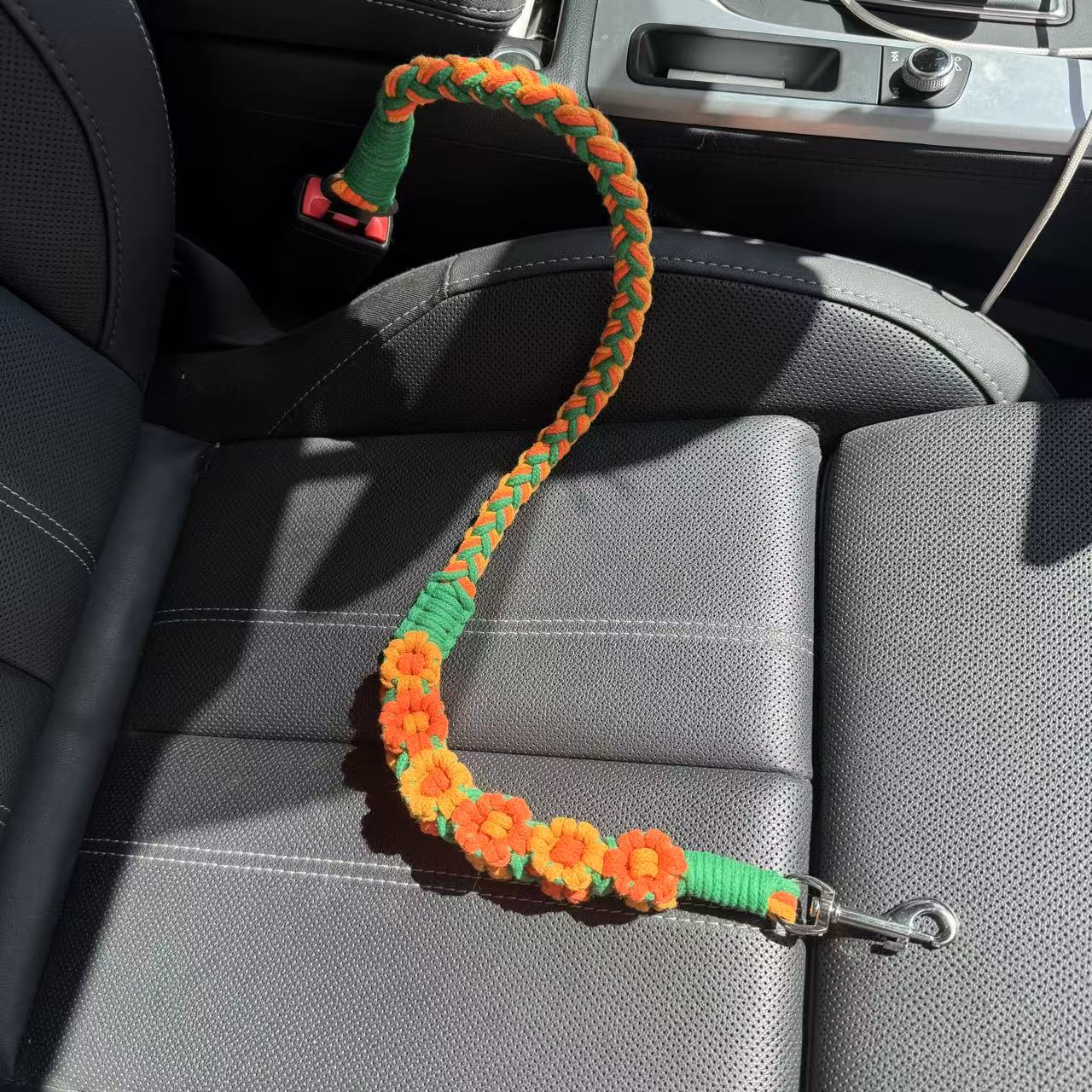 An orange and green handwoven dog safety seatbelt placed on a car seat. The design showcases intricate floral patterns and a secure metal clip, blending functionality with elegance for your pet's travels.