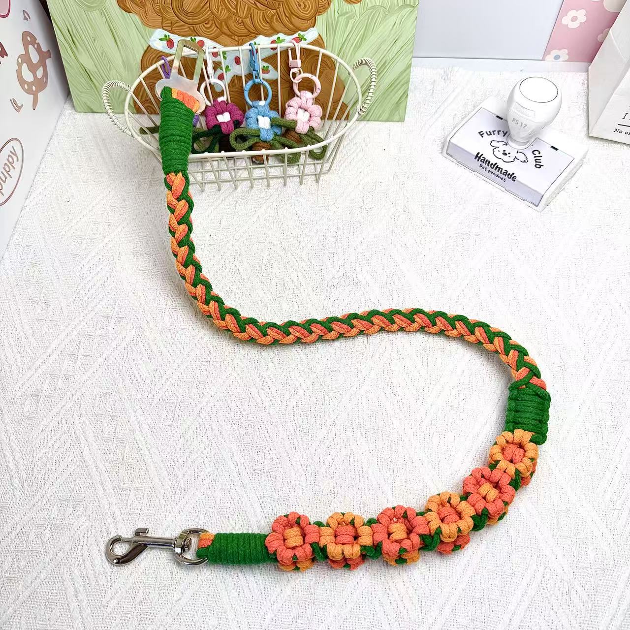 A handwoven orange and green dog safety seatbelt displayed stylishly on a textured surface with floral details and a secure clasp, adding charm to pet accessories.