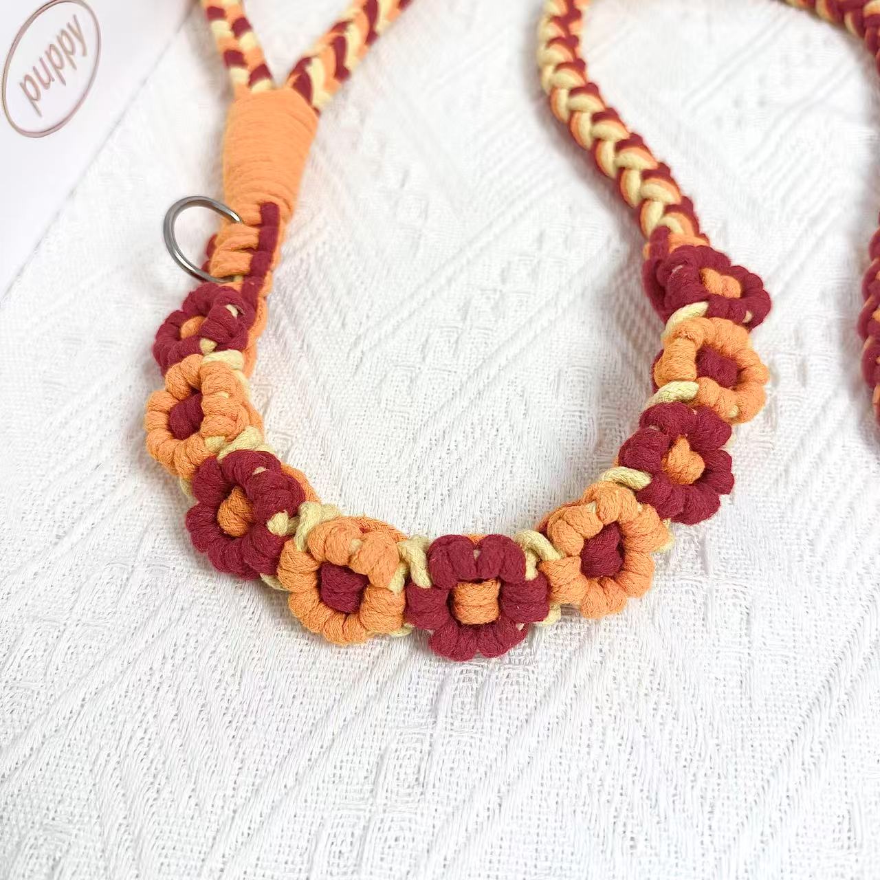 A detailed view of an orange and red handwoven dog leash with a charming floral design. The leash is elegantly placed on a textured white surface, highlighting its intricate craftsmanship and vibrant colors.