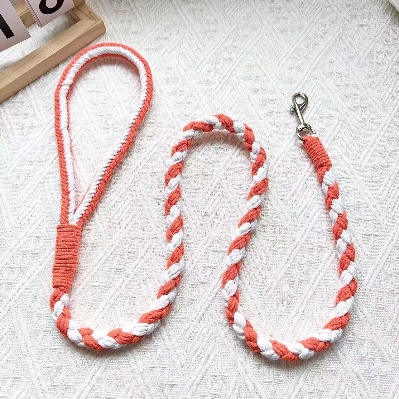 A handmade dog leash in orange and white, featuring a metal clasp, placed on a textured white fabric background.