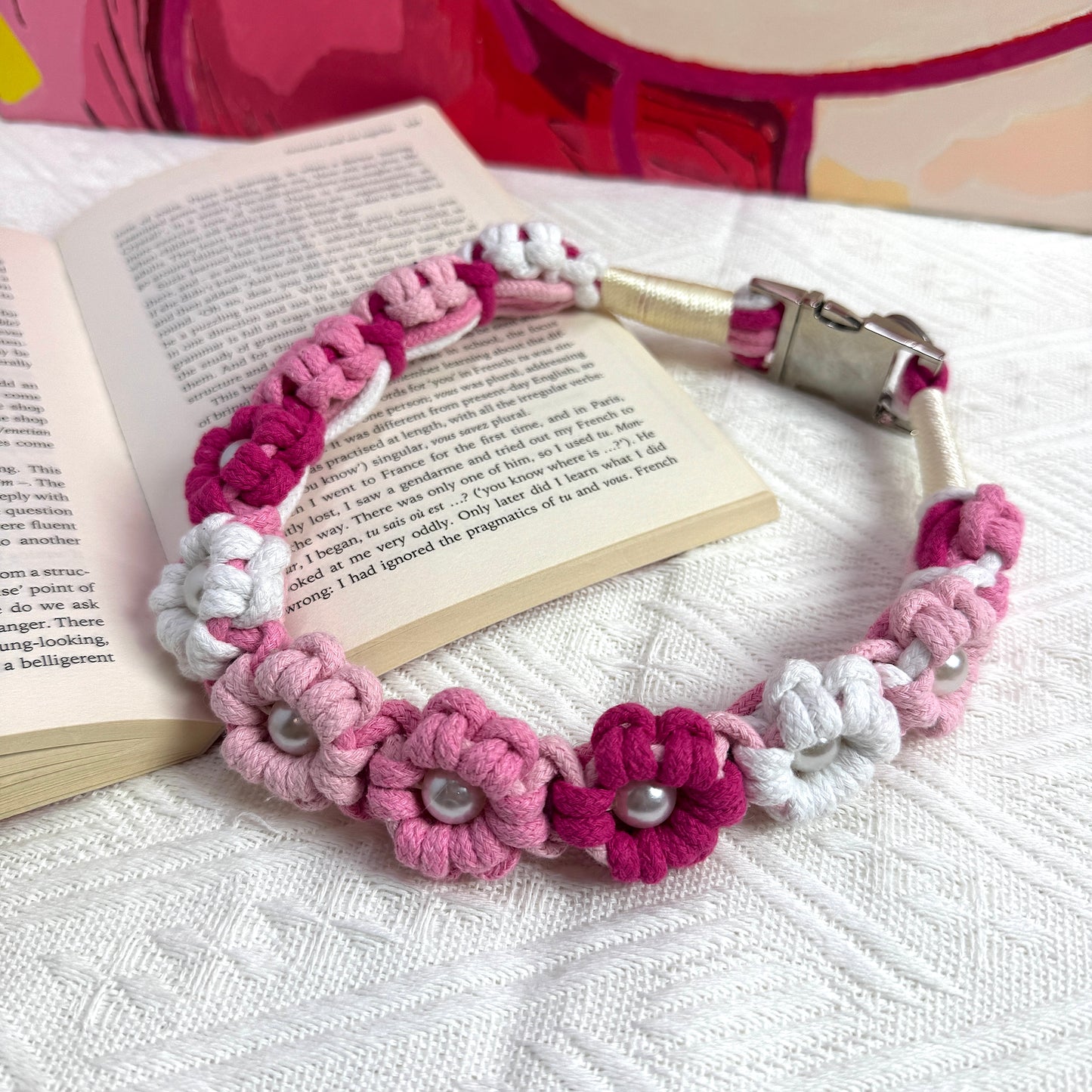 A pink and white handmade dog collar featuring braided flower patterns with pearl embellishments, placed on an open book for a stylish and cozy setting.