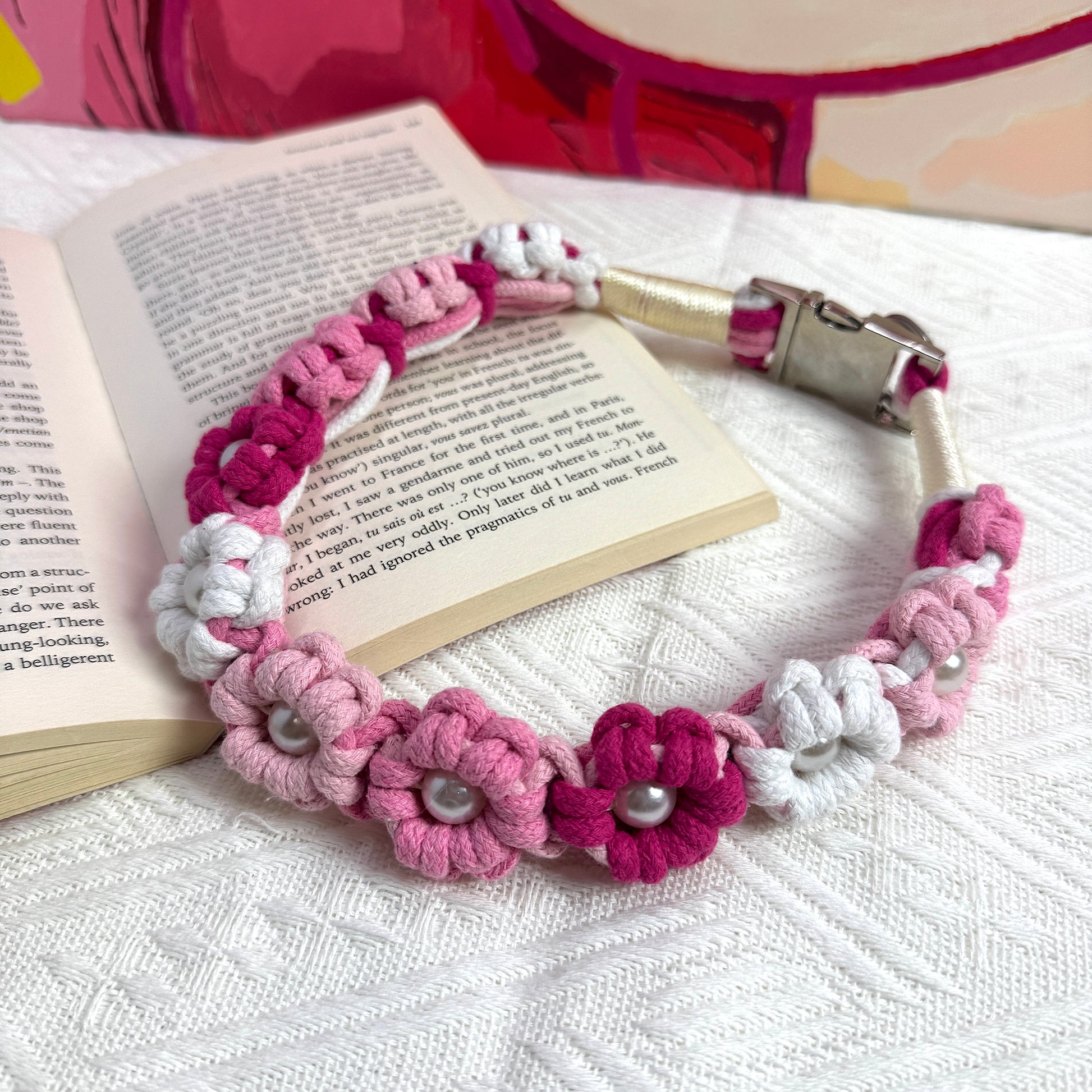 A pink and white handmade dog collar featuring braided flower patterns with pearl embellishments, placed on an open book for a stylish and cozy setting.