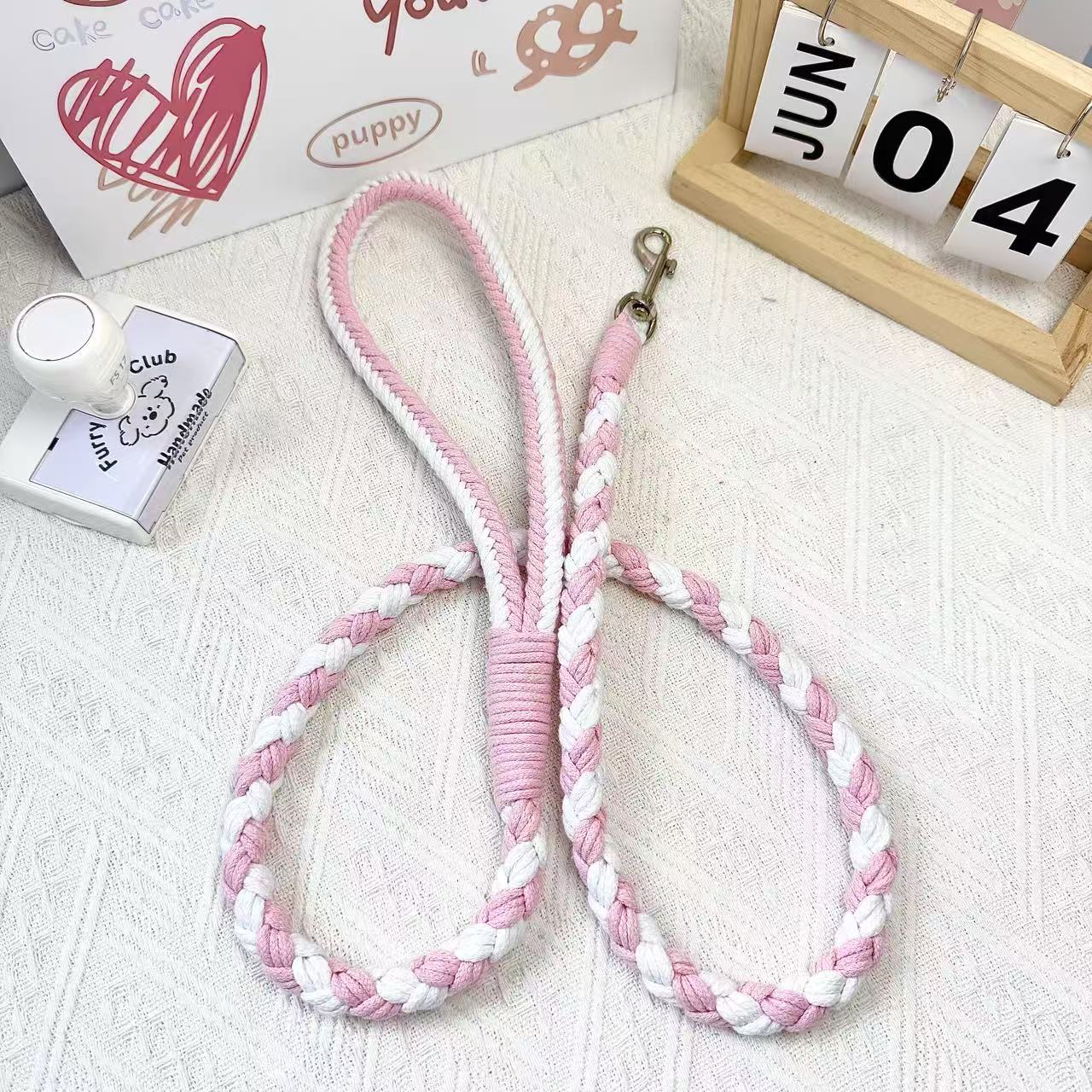 A handmade dog leash in pink and white, featuring a metal clasp, placed on a textured white fabric background.