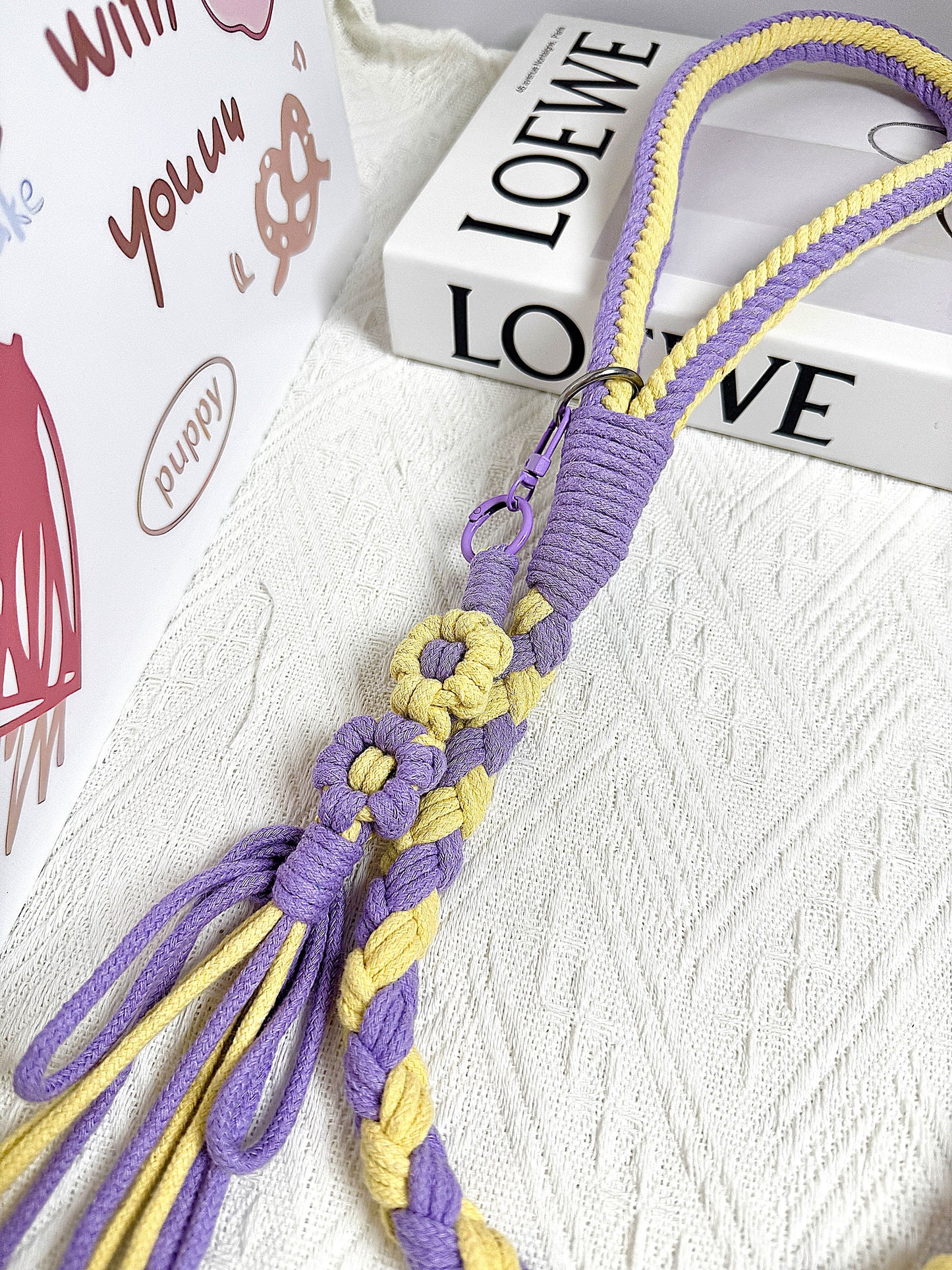 Handmade purple and yellow dog leash with a braided handle and flower charm, styled with a designer book in the background.