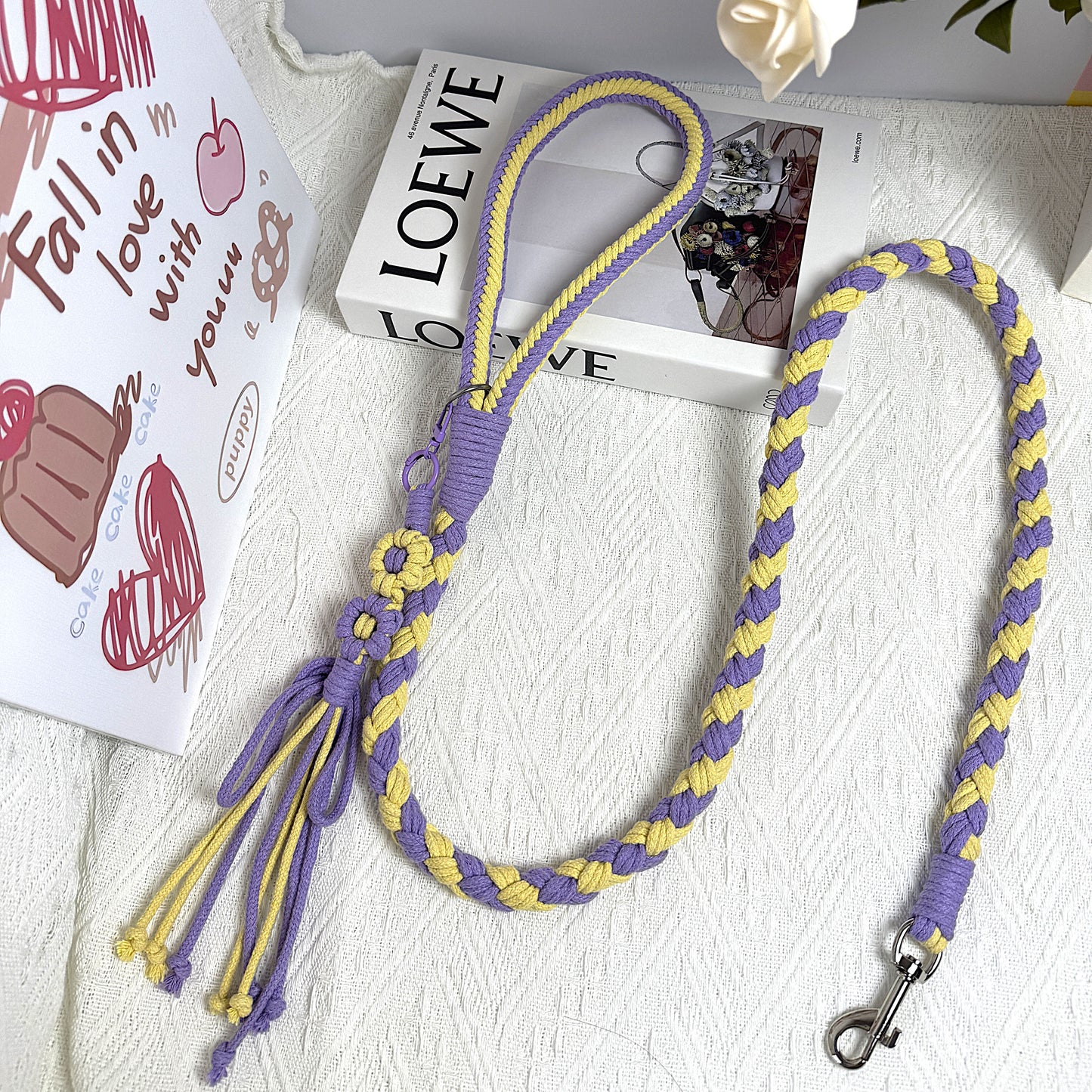 A handmade dog leash in vibrant purple and yellow tones, featuring braided patterns with flower details and tassel ends, elegantly displayed alongside a designer book and decorative background.