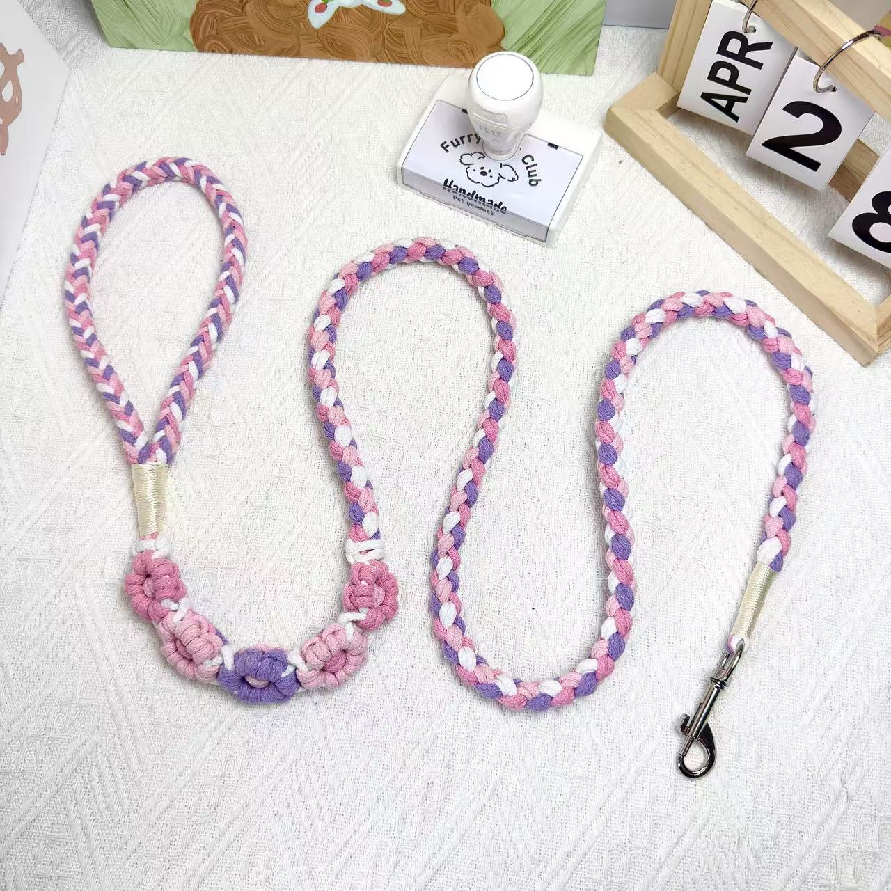 A handmade dog leash in purple and pink with floral accents, featuring a metal clasp, placed on a textured white fabric background.