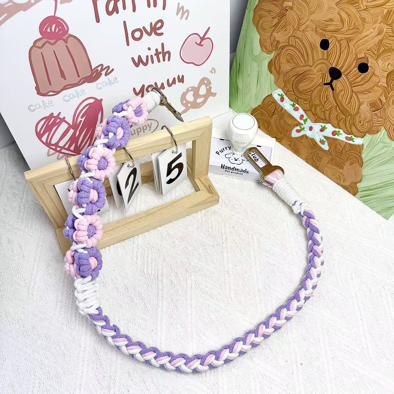 A handmade dog car safety belt in purple and pink with floral accents, featuring a metal clasp and a buckle, placed on a textured white fabric background.