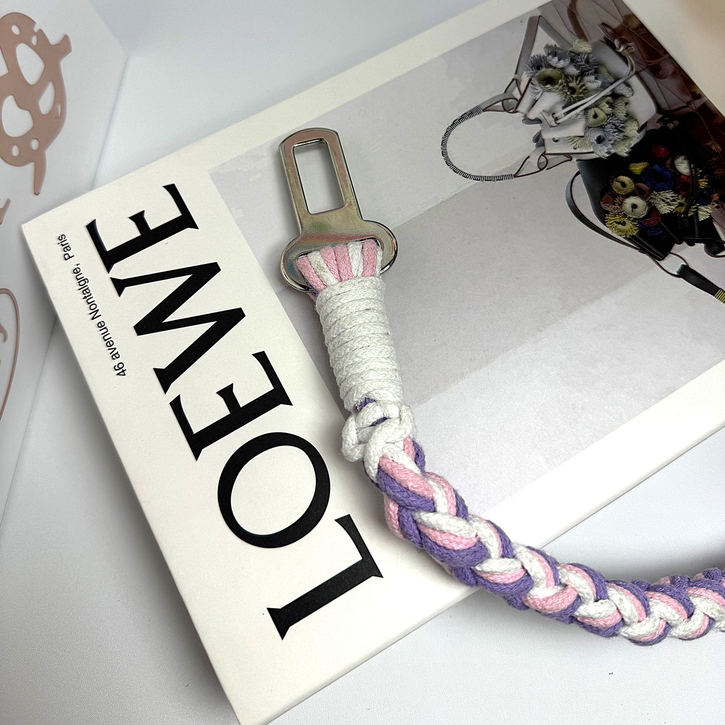 A handcrafted dog seatbelt attachment featuring a braided rope design in pastel pink, purple, and white. The sturdy metal clip is securely wrapped with white cord, ensuring durability.