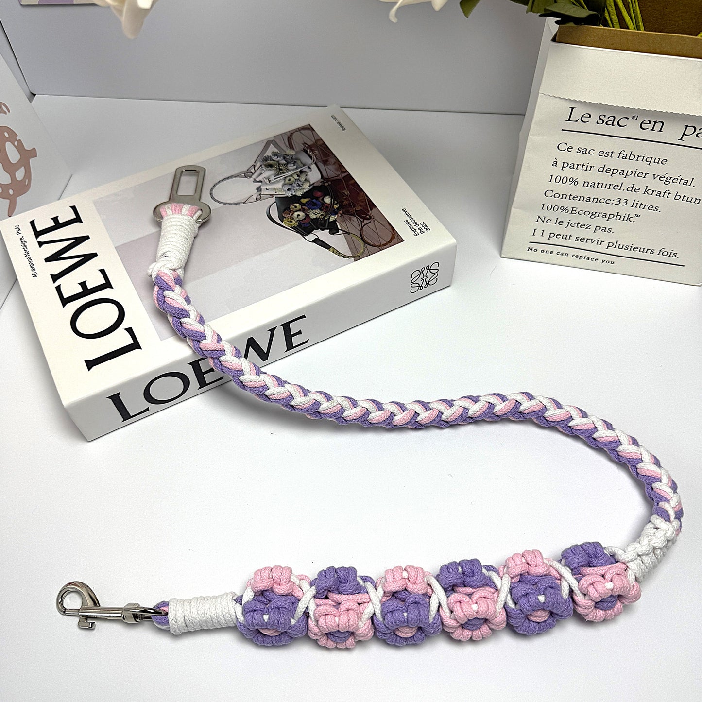 A handwoven purple, pink, and white dog safety seatbelt displayed stylishly on a textured surface with floral details and a secure clasp, adding charm to pet accessories.