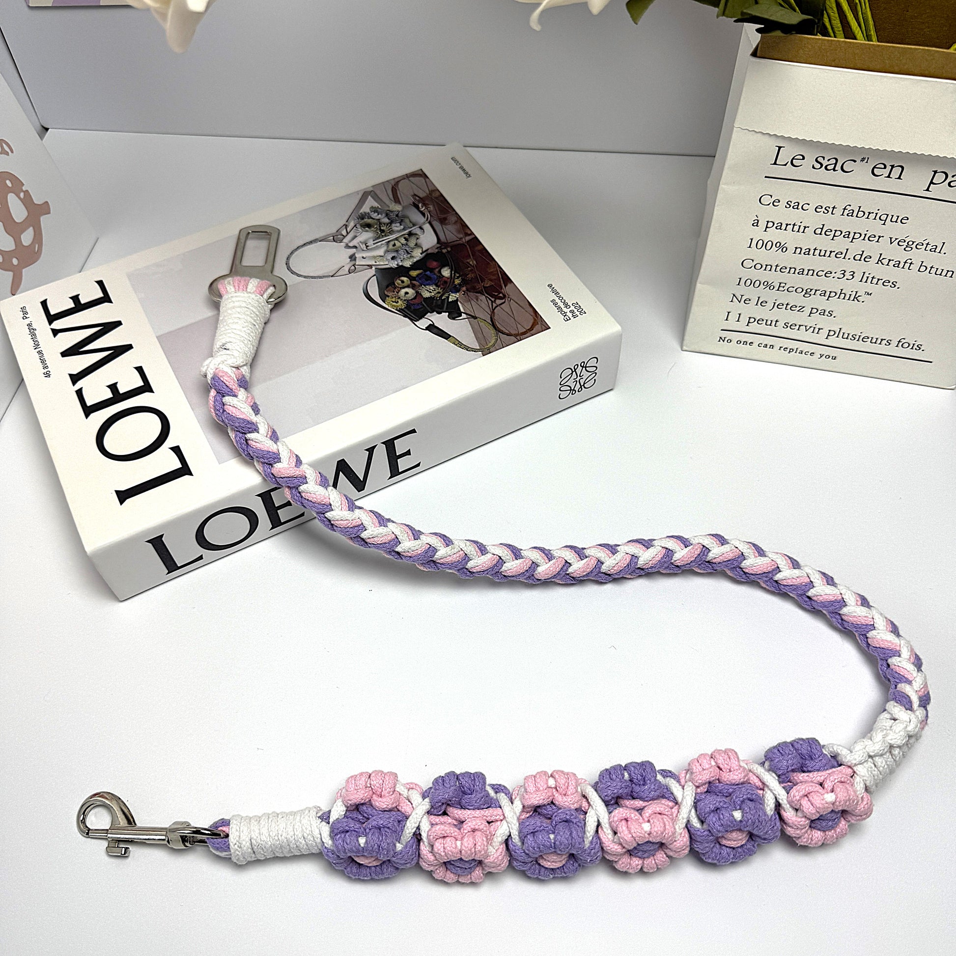 A handwoven purple, pink, and white dog safety seatbelt displayed stylishly on a textured surface with floral details and a secure clasp, adding charm to pet accessories.