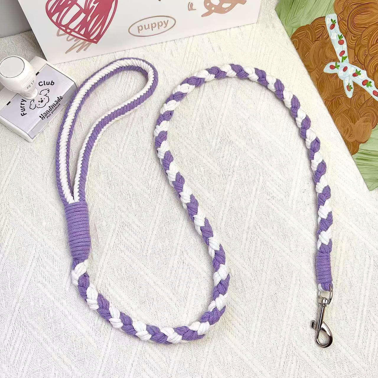 A handmade dog leash in purple and white, featuring a metal clasp, placed on a textured white fabric background.