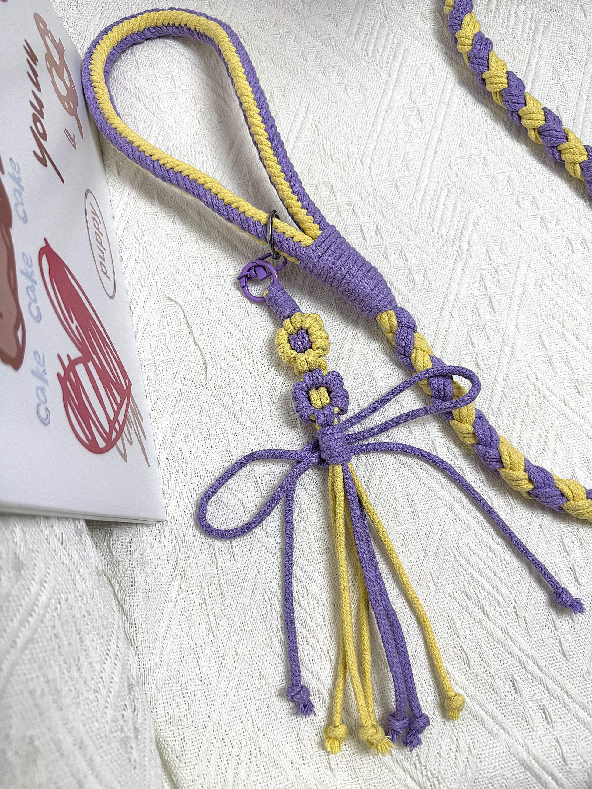 A close-up of a handmade dog leash in purple and yellow, showcasing its comfortable braided handle and decorative flower charm.