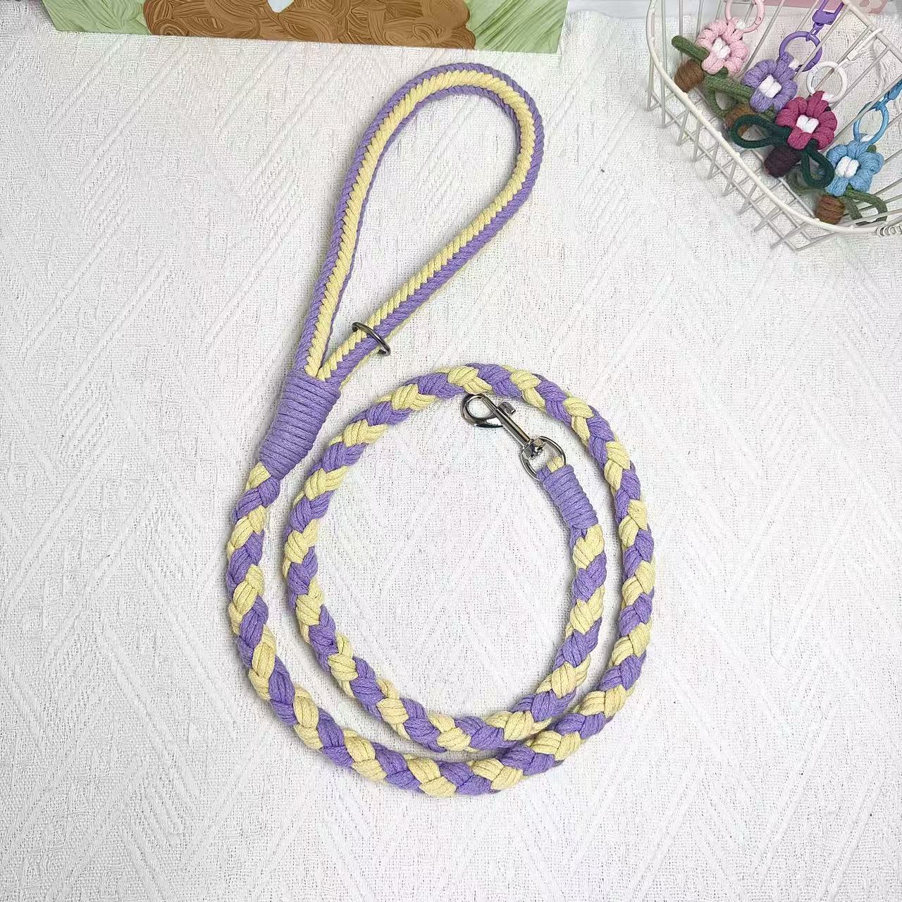 A handmade dog leash in purple and yellow, featuring a metal clasp, placed on a textured white fabric background.