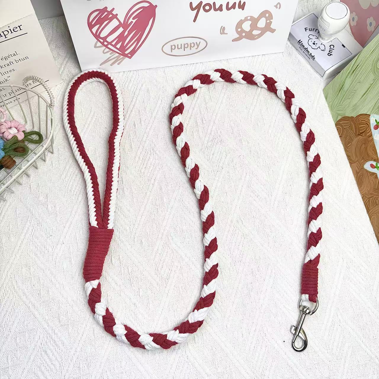 A handmade dog leash in red and white, featuring a metal clasp, placed on a textured white fabric background.