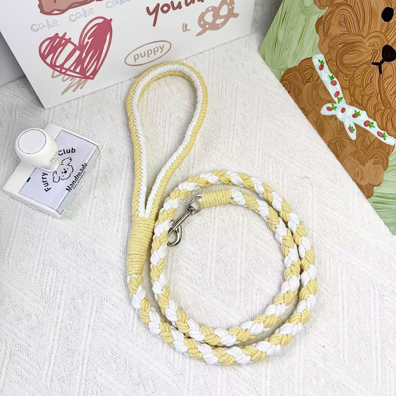 A handmade dog leash in yellow and white, featuring a metal clasp, placed on a textured white fabric background.