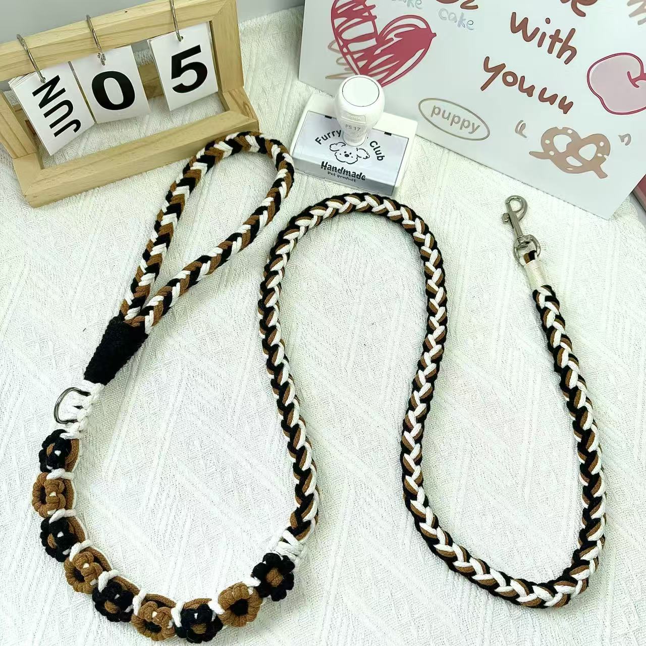 A handmade dog leash in black and brown with floral accents, featuring a metal clasp, placed on a textured white fabric background.