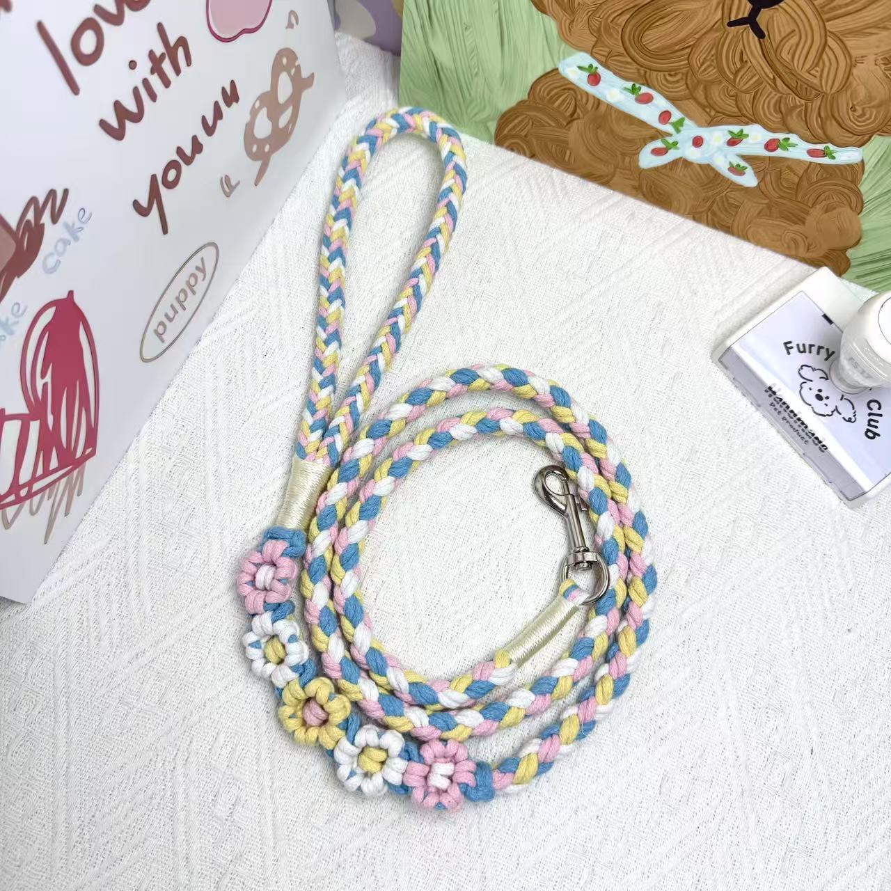 A handmade dog leash in blue and pink with floral accents, featuring a metal clasp, placed on a textured white fabric background.