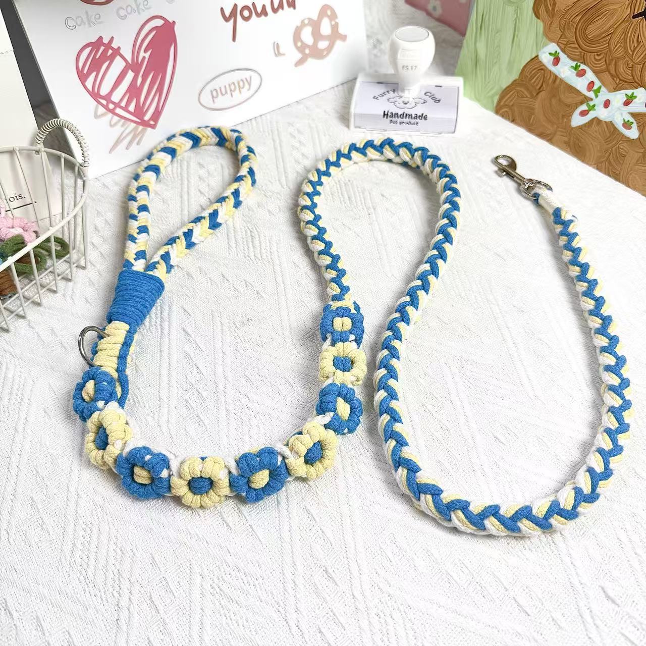 A handmade dog leash in blue and yellow with floral accents, featuring a metal clasp, placed on a textured white fabric background.
