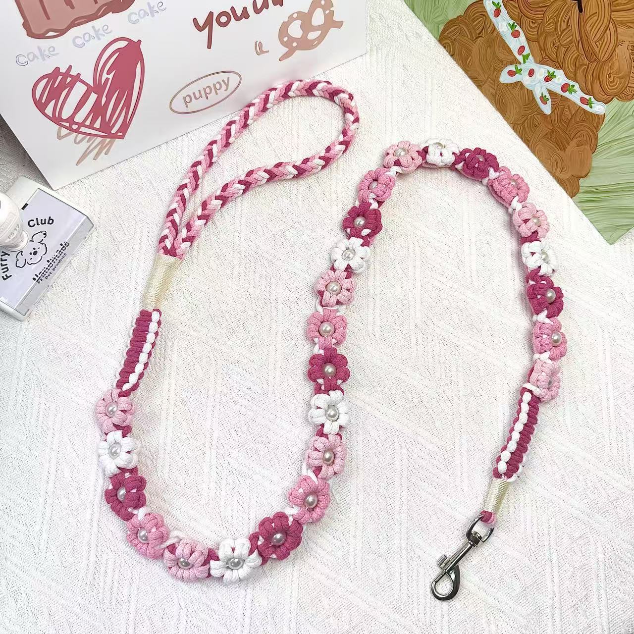 A handmade dog leash in gradient pink with floral accents and pearl details, featuring a metal clasp, placed on a textured white fabric background.