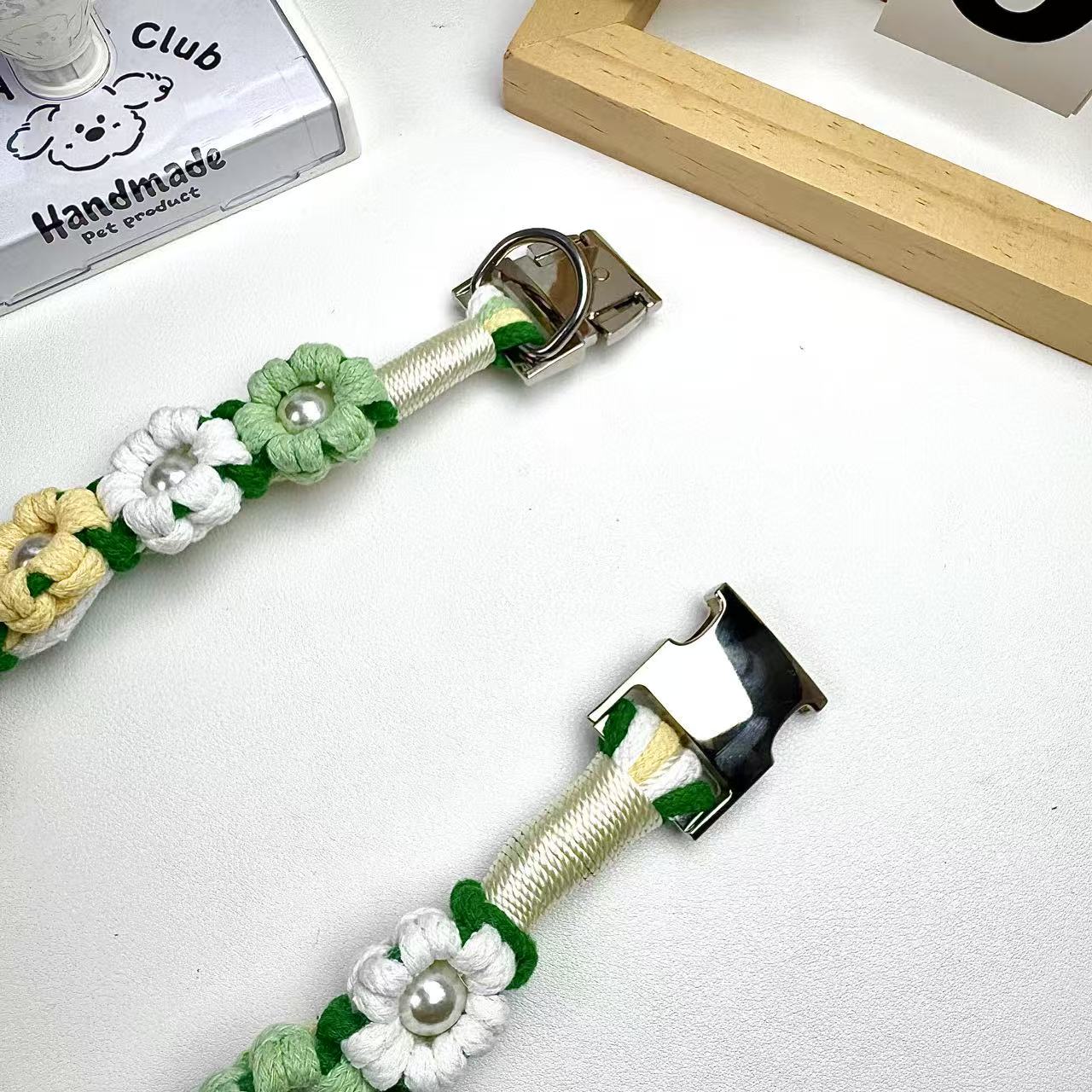 Close-up of a handmade dog collar in green, white, and yellow with flower-shaped knots and pearl centers, featuring a metal clasp on a white background.