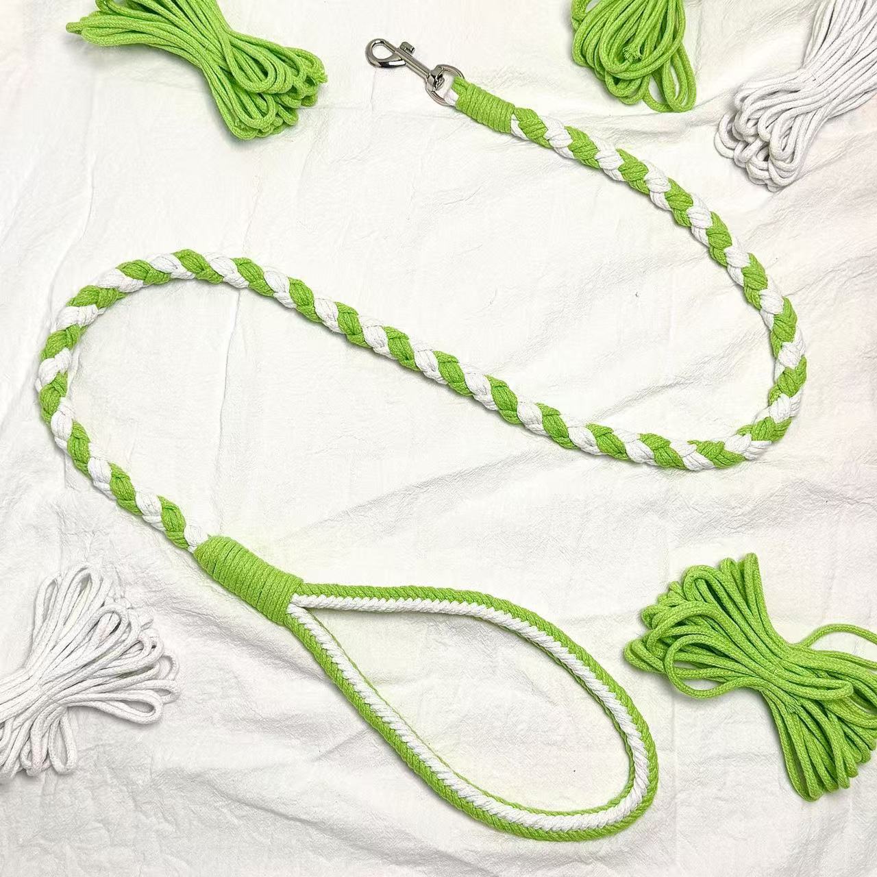 A handmade dog leash in green and white, featuring a metal clasp, placed on a textured white fabric background.