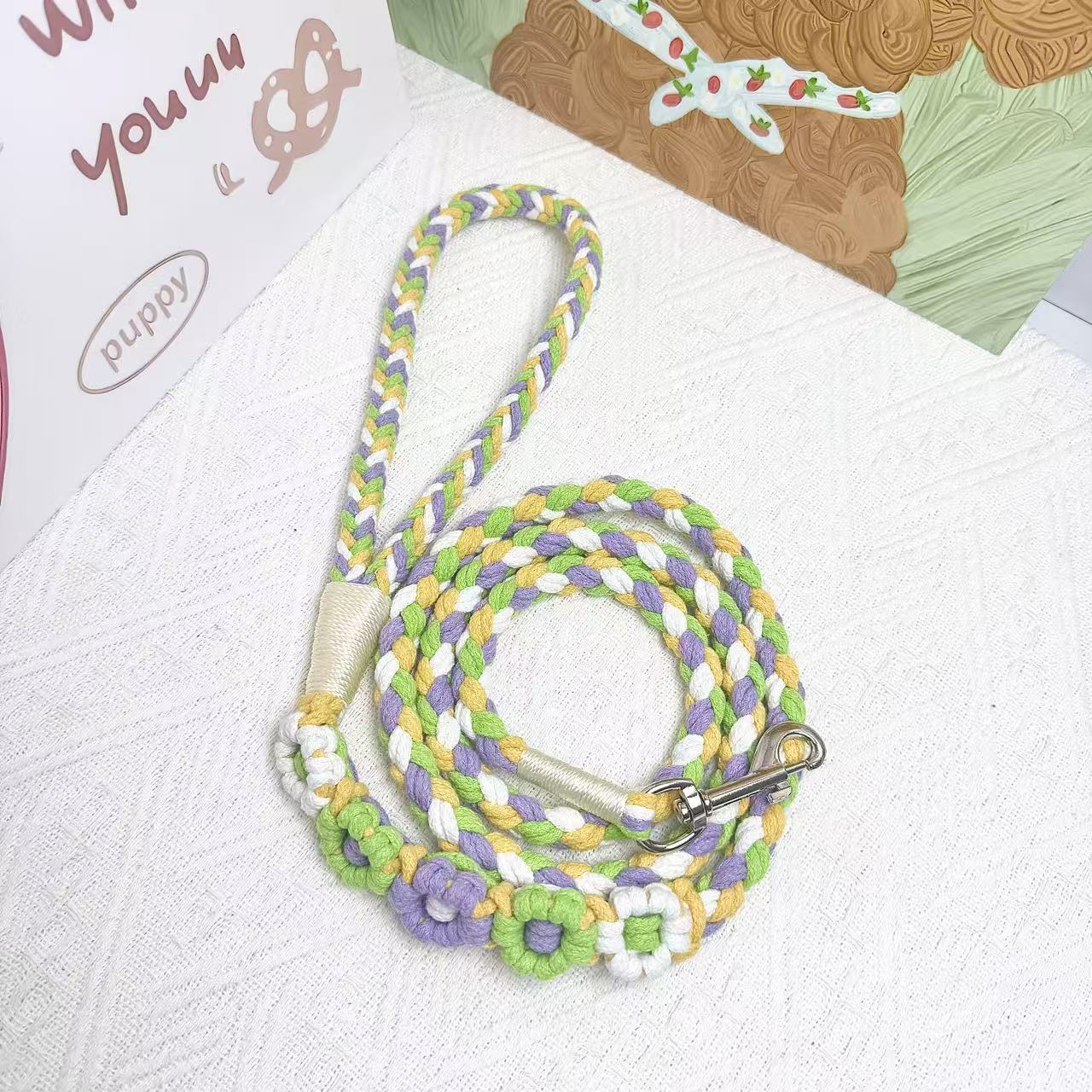 A handmade dog leash in green and yellow with floral accents, featuring a metal clasp, placed on a textured white fabric background.