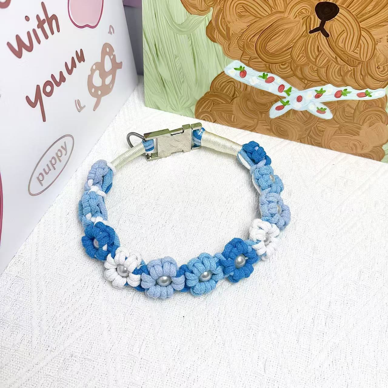 A handmade dog collar in ocean blue with floral accents and pearl details, featuring a metal buckle, placed on a textured white fabric background.
