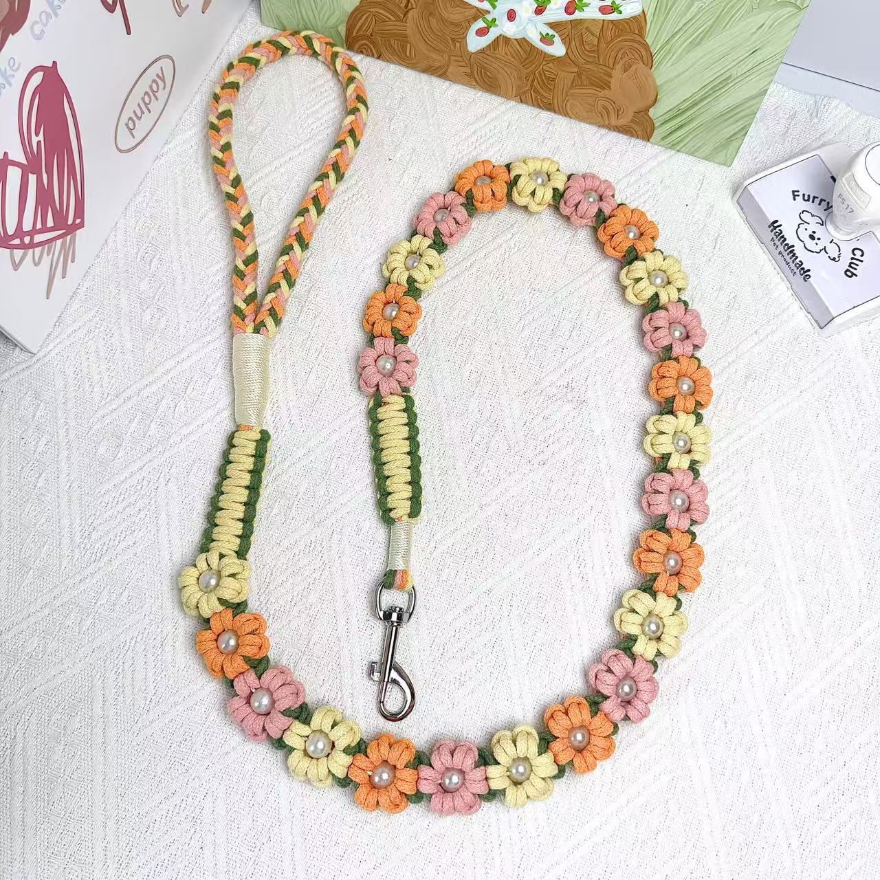 A handmade dog leash in orange and yellow with floral accents and pearl details, featuring a metal clasp, placed on a textured white fabric background.